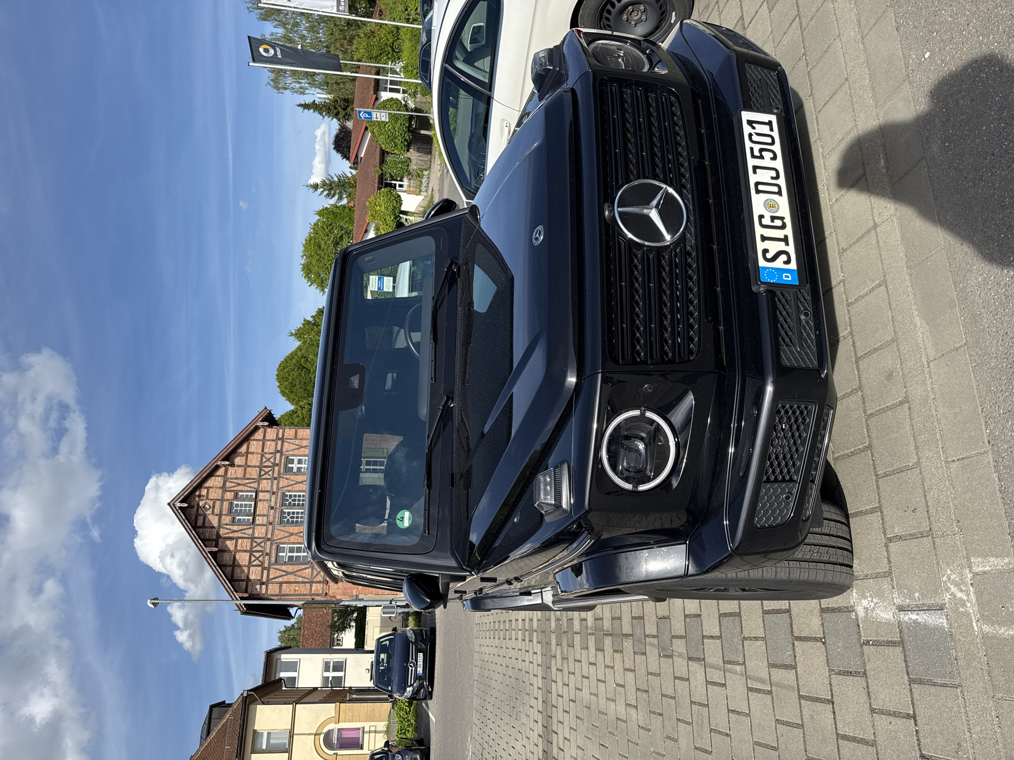The image depicts the front of a black Mercedes-Benz G-Class parked on a brick-paved surface. The vehicle's license plate reads "SIG DJ 501," indicating registration in Sigmaringen, Germany. Behind the car, there is a timber-framed building, partially obscured by a large cloud, suggesting the location is likely in the city of Riedlingen. A parking area, potentially associated with a car dealership or business, surrounds the vehicle. There are other cars parked nearby. The sky is bright blue with scattered clouds, indicating a clear day. The image captures the car in an outdoor setting in the town of Riedlingen.