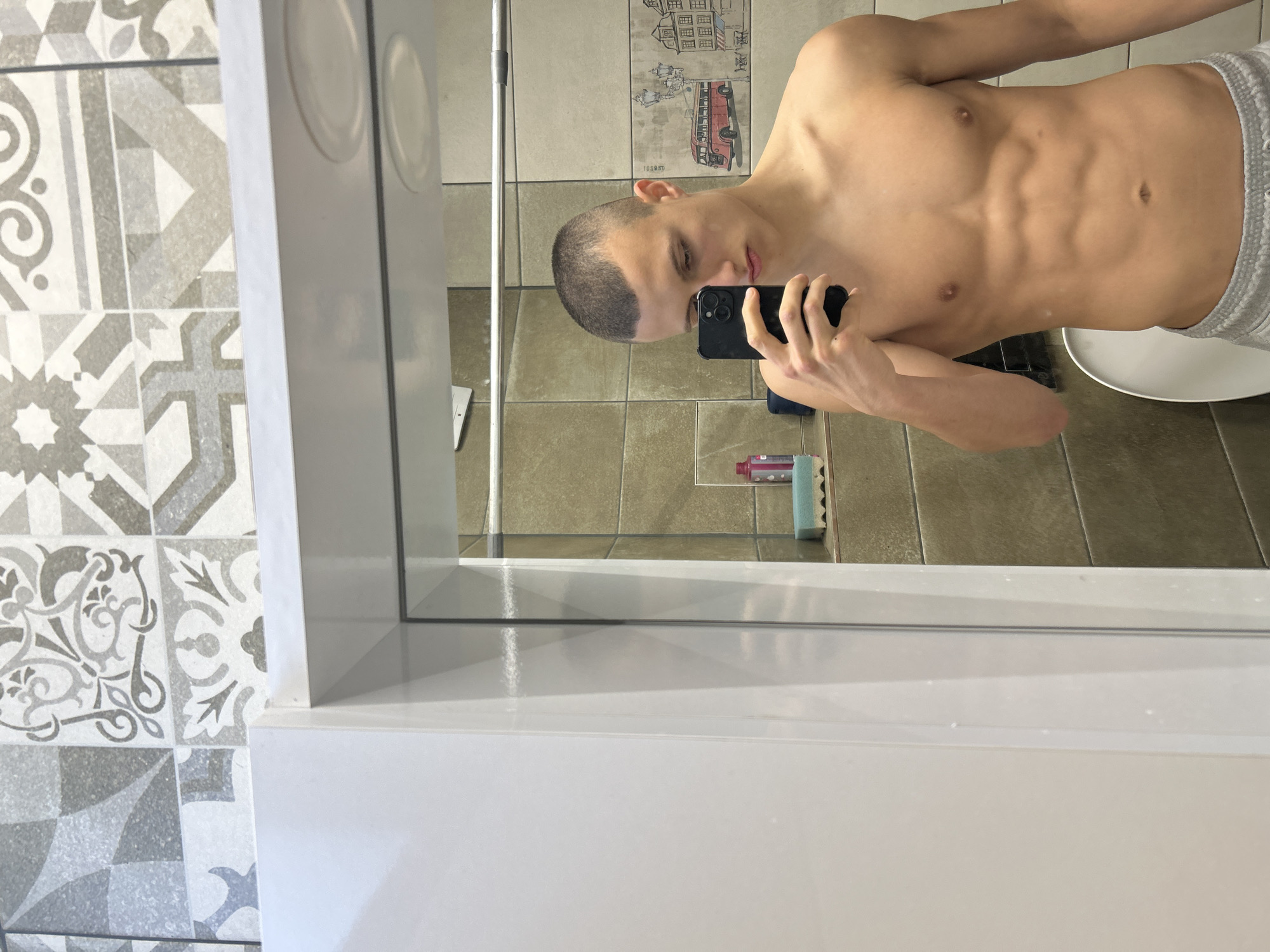 A shirtless, light-skinned young man with a closely shaved haircut is taking a selfie in a bathroom mirror. He is holding a black smartphone in his right hand. His abdomen is visible. The bathroom has tiled walls and floor. A toilet is partially visible. There's cleaning supplies on the floor beneath the mirror. The location appears to be Burgas, Bulgaria.