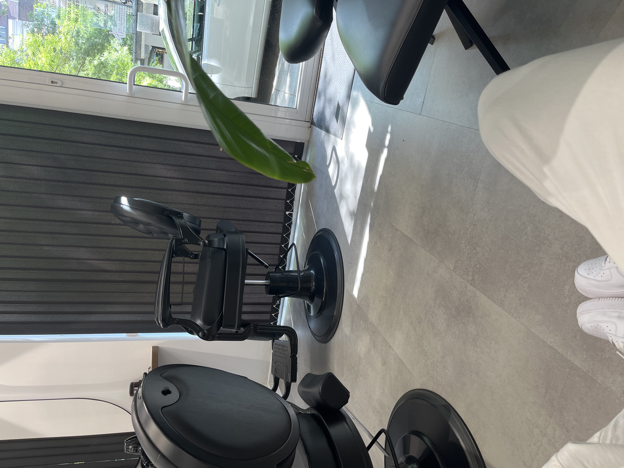 The image depicts the interior of a commercial establishment, likely a barbershop or hair salon, situated in the city of Sofia, Bulgaria. The perspective is low, originating from a seated individual whose lower legs, clad in white trousers, and white athletic shoes (Nike Air Force 1s) are visible in the foreground. A blurred green plant leaf occupies the immediate foreground.
The floor is covered with large, light gray, mottled tiles. In the midground, two black, hydraulic barber chairs are prominent. To the left, the back and an armrest of one chair are partially visible. In the center-right, a full classic-style barber chair with prominent armrests, a footrest, and a circular base is positioned, facing away from the viewer. A third black chair, partially visible on the far right, suggests additional seating.
The left wall is white. On the right, a large window or glass door with a white frame allows significant natural light into the space, casting distinct shadows on the floor. Dark gray vertical blinds partially cover this window/door. A black doormat is visible on the floor near the threshold of the entrance.
Beyond the window/door, the exterior reveals green trees with full foliage, indicating a daytime setting. A white car, possibly an Audi based on a faint reflection, is parked outside. Text in Bulgarian is visible, mirrored, on the window. Identifiable words include "ТАИР" (TAIR), operating hours "09:00 - 18:00" and "09:00 - 13:00", days "ПОНЕДЕЛНИК-ПЕТЪК" (Monday-Friday) and "СЪБОТА" (Saturday), and labels such as "ПОЧИВКА" (Rest/Closed), "ПРОИЗВОДСТВО" (Production), "ИНДУСТРИАЛНА ЗОНА" (Industrial Zone), "ЦВЯТ" (Color), and "ПРОДАЖБИ" (Sales). A QR code is also visible on the window. These Bulgarian language details confirm the location in Sofia, Bulgaria, within an area designated as an "Industrial Zone."