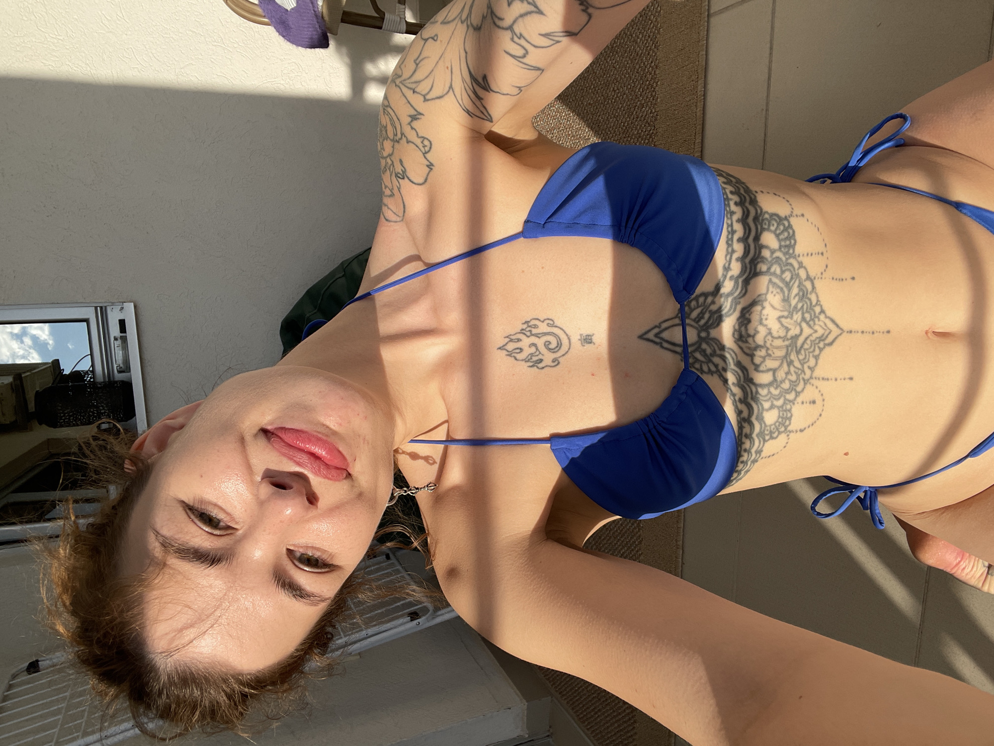A young woman is posing for a selfie while sunbathing outdoors. She is wearing a blue bikini. She has multiple tattoos on her arms, chest, and abdomen. The location appears to be a balcony or patio in Rafailovići, Montenegro, based on the building style and likely sunny climate. There is a view of the sky and adjacent buildings through a window frame. The ground covering seems to be a woven mat.