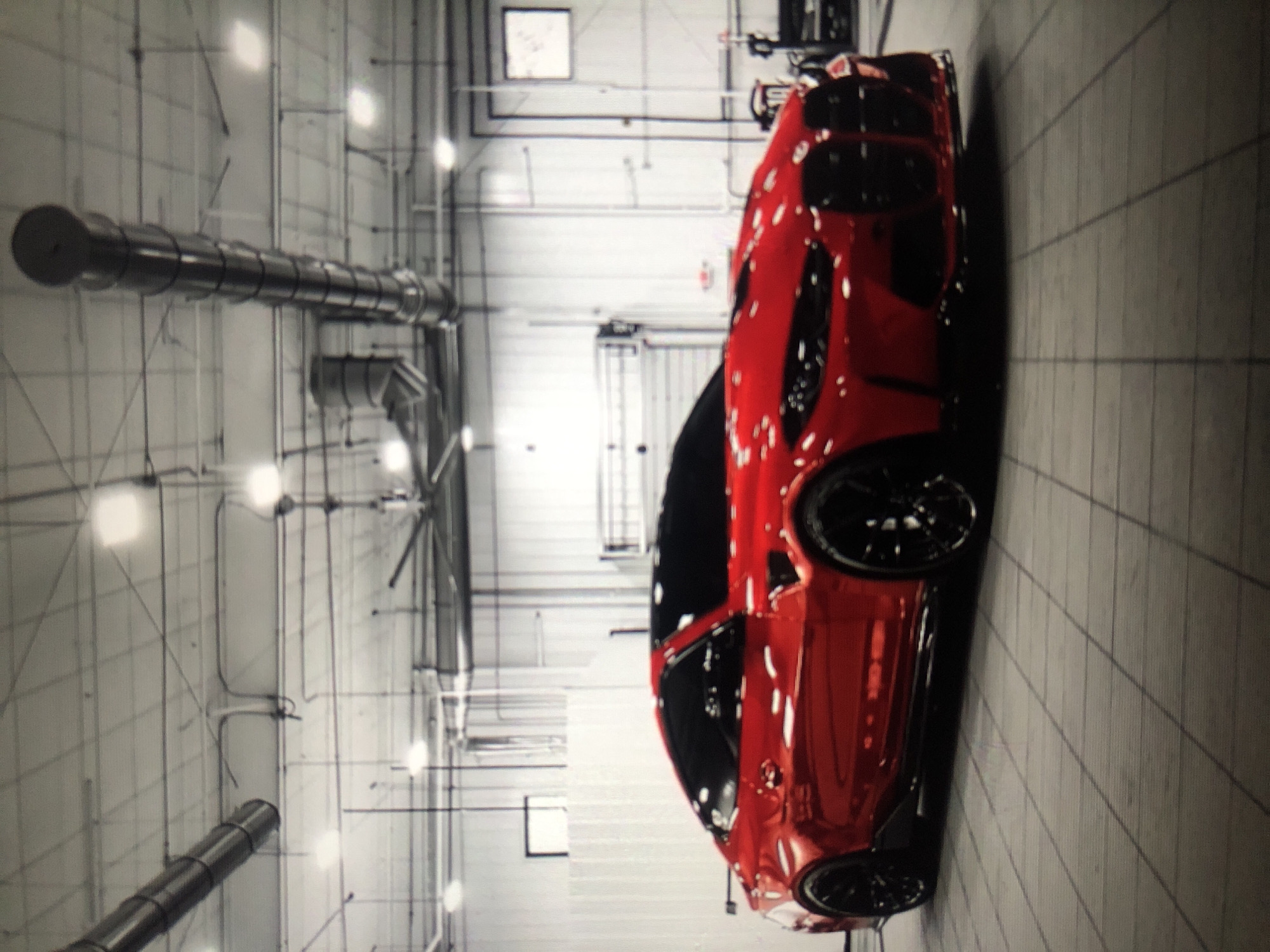 A red car is parked in what appears to be a warehouse or industrial-looking space. The walls and ceiling are a light color and feature a grid-like pattern of pipes and conduits, as well as industrial lighting fixtures. The floor is tiled. There are no people visible in the frame. The specific location in Lhokseumawe, Indonesia, cannot be determined from the visual information provided.