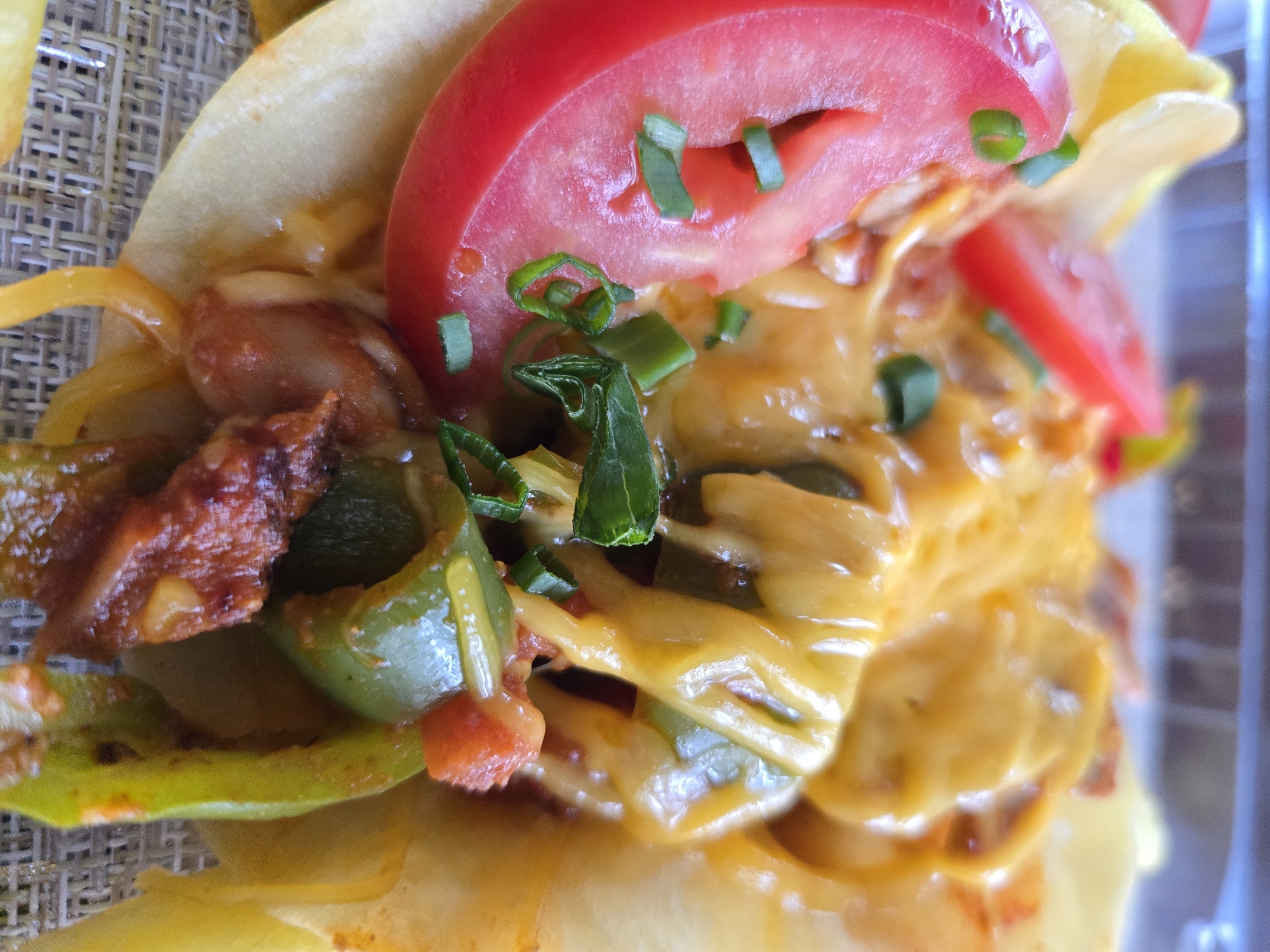 A close-up shot captures a partially obscured taco, likely situated in an unknown location, possibly a restaurant or food stall. The taco features a yellow tortilla, adorned with cooked peppers (both green and orange), cooked meat, melted yellow cheese, slices of red tomato, and garnished with chopped green scallions. The taco is presented on a textured, woven surface.