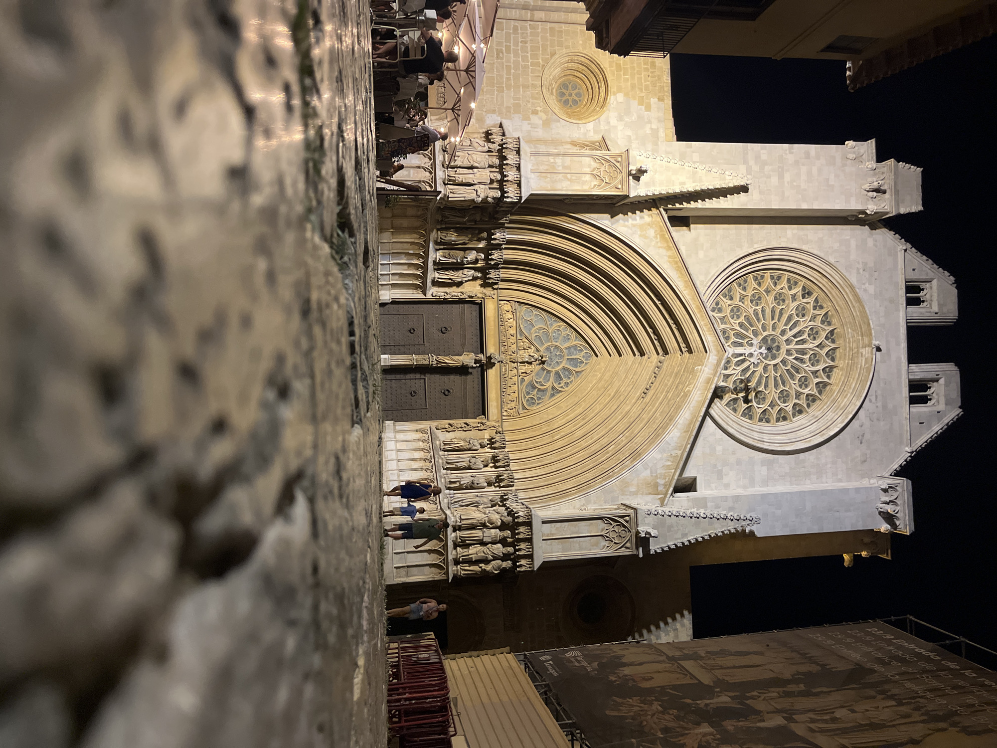A low-angle shot captures the facade of the Tarragona Cathedral at night, with the foreground dominated by a close-up of the cobblestone street. The cathedral's illuminated Gothic architecture features a large rose window above a grand arched entrance adorned with numerous sculptures. To the left, an outdoor seating area with umbrellas and people dining is visible, suggesting a restaurant or cafe. On the right, a tall banner with text and images is partially visible. Two adults and a child are walking towards the cathedral entrance, while a woman stands to their right. Other people are seen in the background, both at the dining area and near the cathedral. The lighting creates dramatic shadows and highlights, emphasizing the texture of the stone and the night sky.