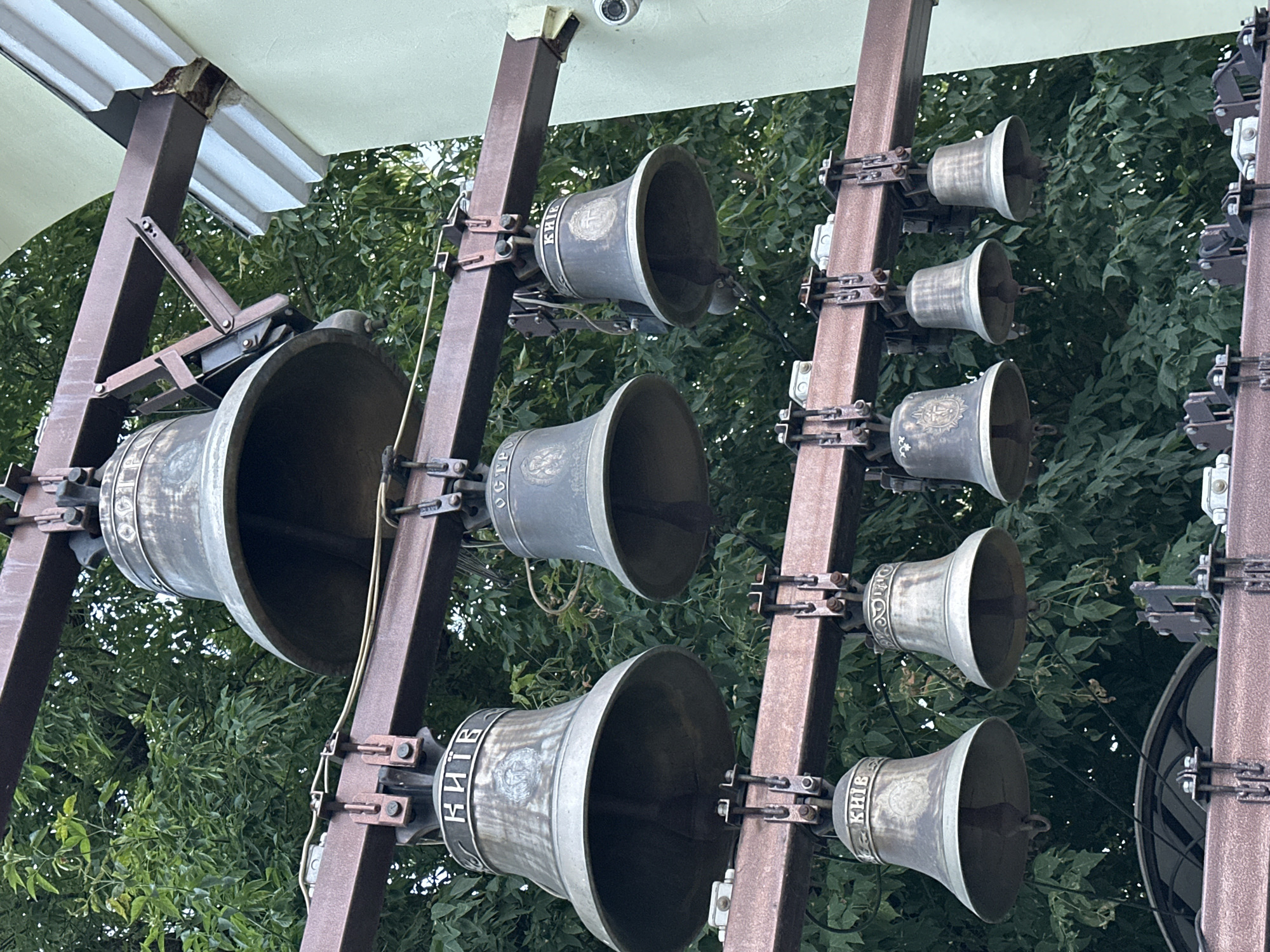 The image shows a close-up view of a set of bells hanging from metal beams, with lush green foliage in the background. The bells are made of bronze and appear to be old, with visible inscriptions. The setting is likely outdoors, possibly part of a church or bell tower in Kyiv, Ukraine, given the inscription "КИЇВ" (Kyiv) on some of the bells. The bells are arranged in multiple rows, with larger bells at the top and smaller ones below. The metal beams are a reddish-brown color and show signs of rust and weathering. A security camera is visible mounted on a light green wall on the right side of the image. The overall impression is one of historical significance and traditional craftsmanship.