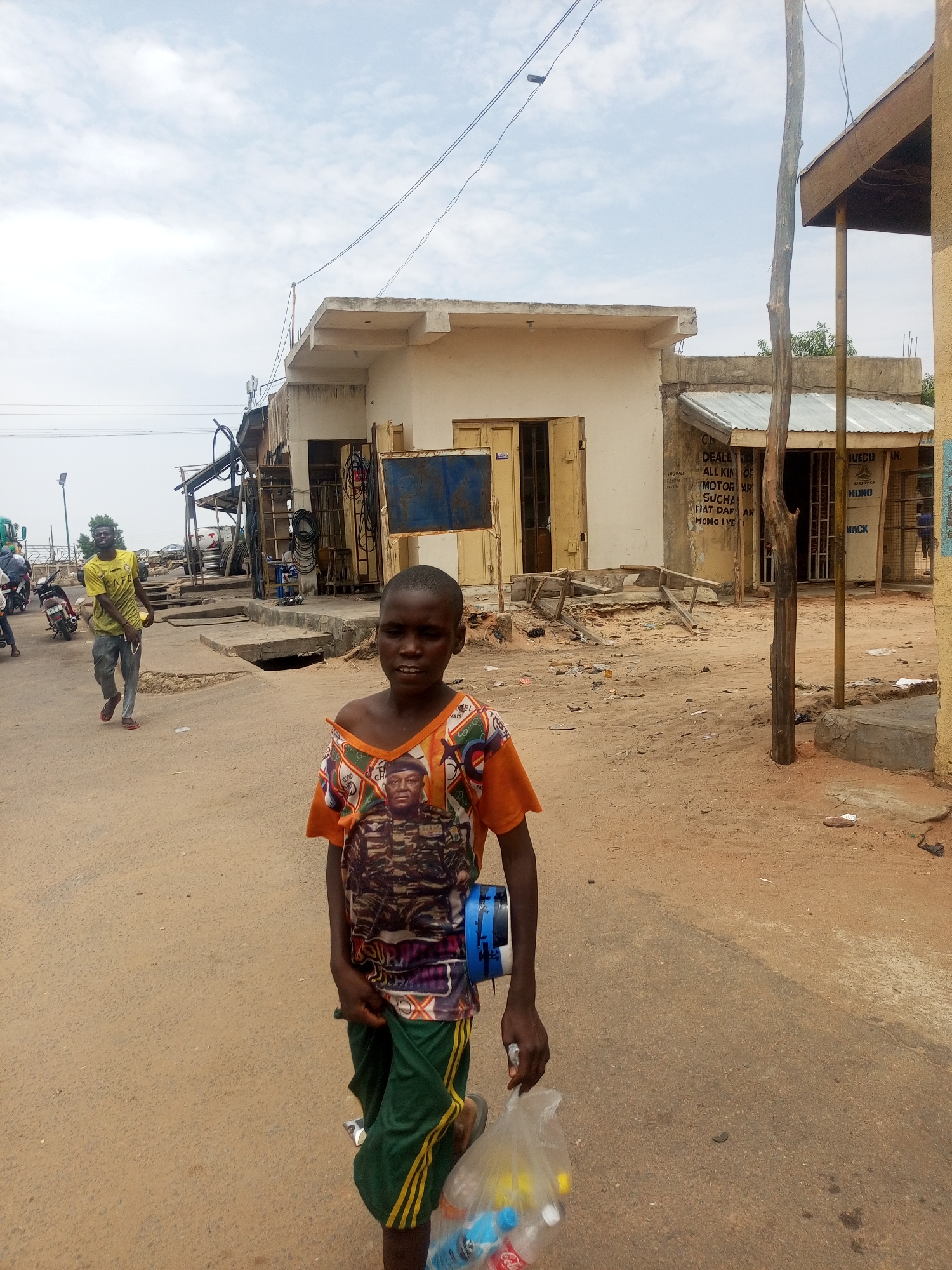 A medium shot captures a young person wearing an orange shirt with a graphic print of a man in military attire. The person is carrying what appears to be a makeshift musical instrument or drum and a plastic bag filled with bottled beverages. They are standing on a dirt road in what appears to be a city street.
In the background, there are buildings with worn facades, and other people are walking on the street, including an individual in a yellow shirt. There are utility lines visible in the sky. One of the buildings has a sign with text that appears to say "DEALE...ALL KIND OF...MOTOR AR..." The area seems to be in a state of disrepair, with debris scattered on the ground. Based on information, this scene takes place in Tempure, Nigeria.