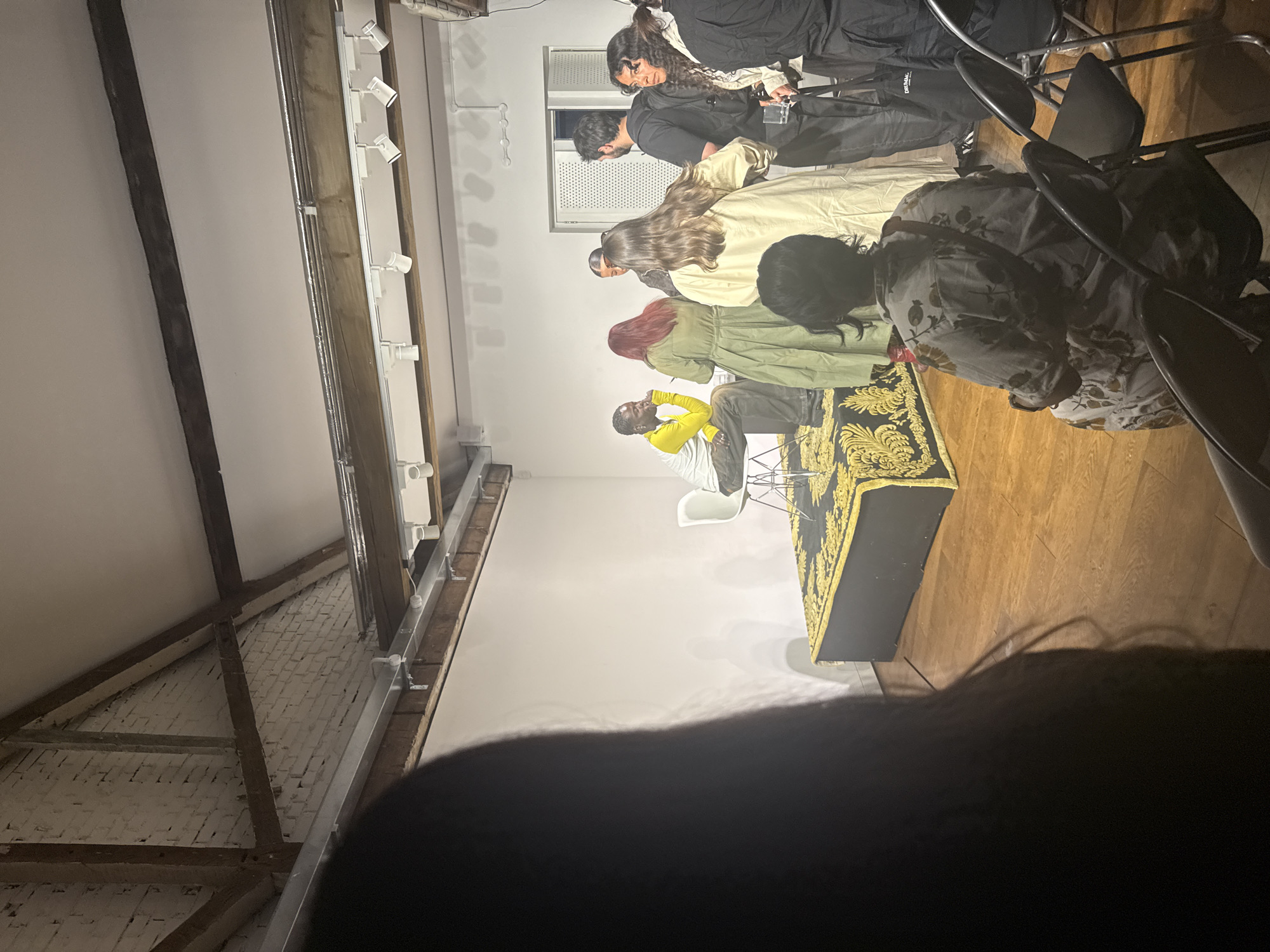 The image captures an indoor gathering, likely a presentation or a talk, within a space characterized by white walls, wooden flooring, and exposed wooden beams and brickwork in the ceiling area. The setting suggests an event space, gallery, or repurposed industrial building.
Several individuals are present. In the center, a person wearing a yellow long-sleeved top and dark pants is seated on a white modern chair, appearing to be the focal point of the event. They are looking upwards. Directly in front of them is a low, rectangular object draped with an ornate black and gold patterned fabric, possibly a speaker's podium or a display table.
A small audience or group of attendees surrounds this central figure. On the left side, facing away from the camera, are several standing individuals: one with red hair in a green dress, another with long brown hair in a light-colored dress, and one with dark hair. Further back and to the right, a person with dark, curly hair and a patterned shirt is standing, looking towards the seated individual. Another person, dressed in black, stands near a window with an air conditioning unit. On the far right, partially visible, is a person seated on a black folding chair, also facing away from the camera. Additional black folding chairs are arranged nearby.
Track lighting fixtures are visible on the ceiling, illuminating the scene. A large, dark, blurry shape in the foreground indicates the photo was taken from behind someone's head or shoulder.
A notable detail is the word "BALENCIAGA" visible on a black bag or piece of clothing in the upper right background. The atmosphere appears attentive and engaged. The overall impression is an organized event taking place in a contemporary yet somewhat rustic venue.