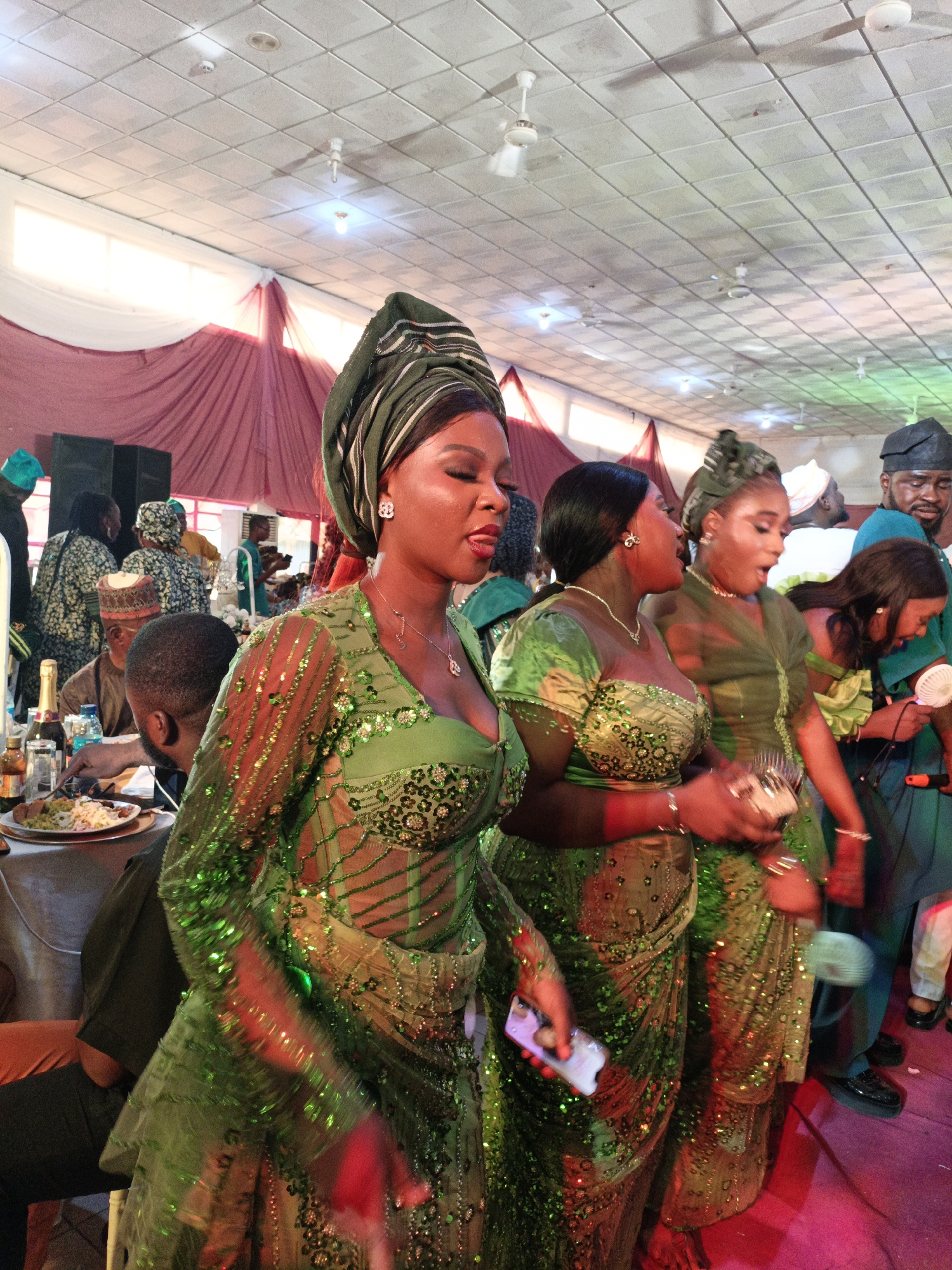 The image captures a celebration in Fagge D2, Nigeria. Three women in similar green, sequined dresses and headwraps are the focal point, seemingly engaged in a lively interaction, possibly singing or dancing. One woman holds a mobile phone. In the background, other individuals are visible, along with tables set for a meal with visible plates, drinks, and what appears to be a bottle of champagne. The setting appears to be indoors in a large hall or event space with ceiling fans and draped fabric decorations.