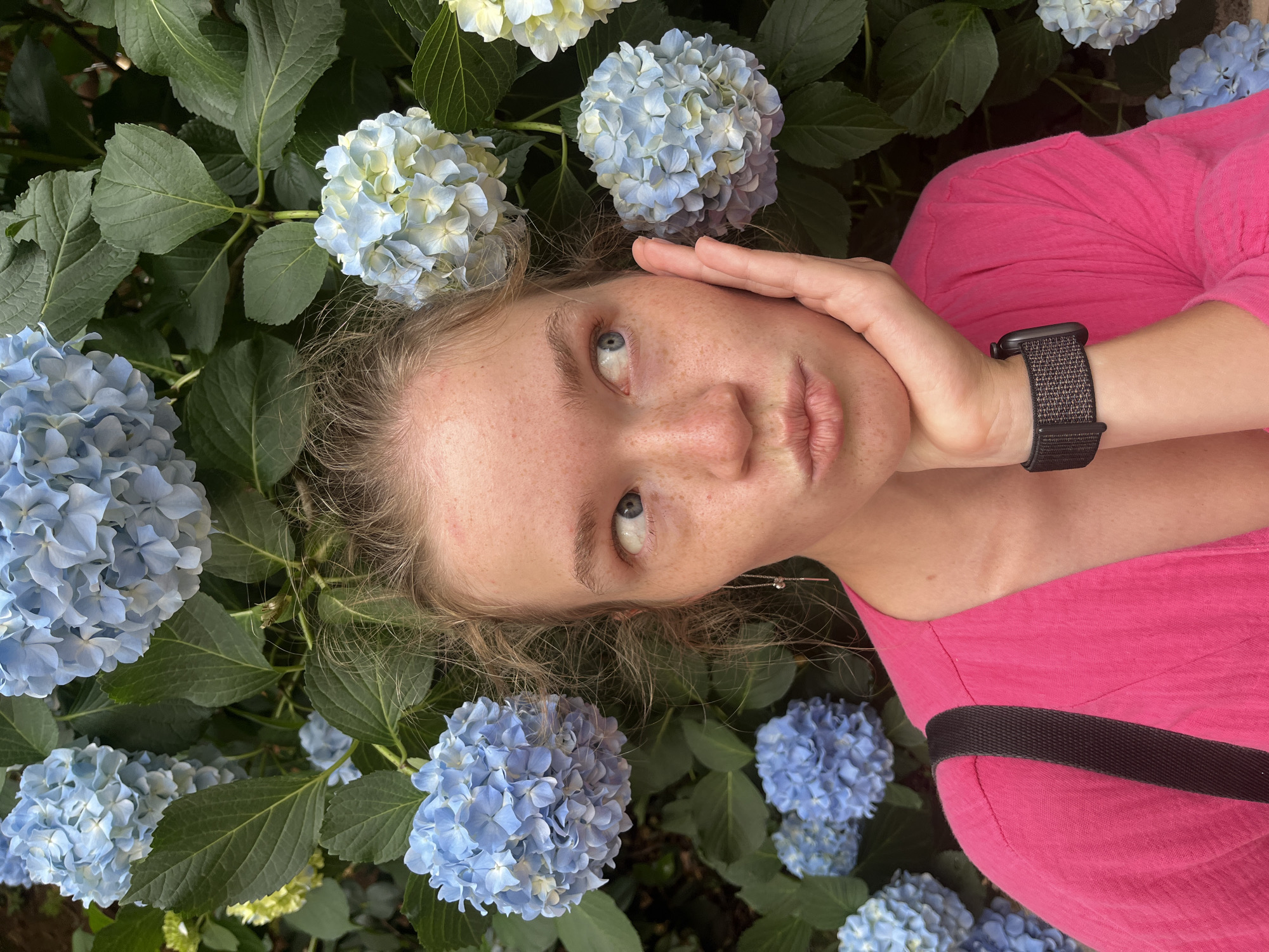 A close-up portrait, rotated clockwise, features a person nestled amongst vibrant blue and light blue hydrangeas. The subject has light brown hair, striking blue eyes, and numerous freckles across their nose and cheeks. They are looking upwards and slightly to the right, with a thoughtful or curious expression. One hand is resting gently on the side of their forehead.
The person is wearing a bright pink t-shirt or top. A dark, woven-strap watch (possibly a smartwatch) is visible on their left wrist, and a dark strap, likely from a bag, crosses their chest. A delicate silver earring with a small drop can be seen near their right ear.
The background is dominated by lush green foliage and clusters of blooming hydrangeas. Some flowers are a rich blue, while others display a mix of pale blue and creamy white petals. The setting appears to be an outdoor garden or bush in Budva, Montenegro, during the daytime, with soft, natural light illuminating the scene. No text is visible in the image.