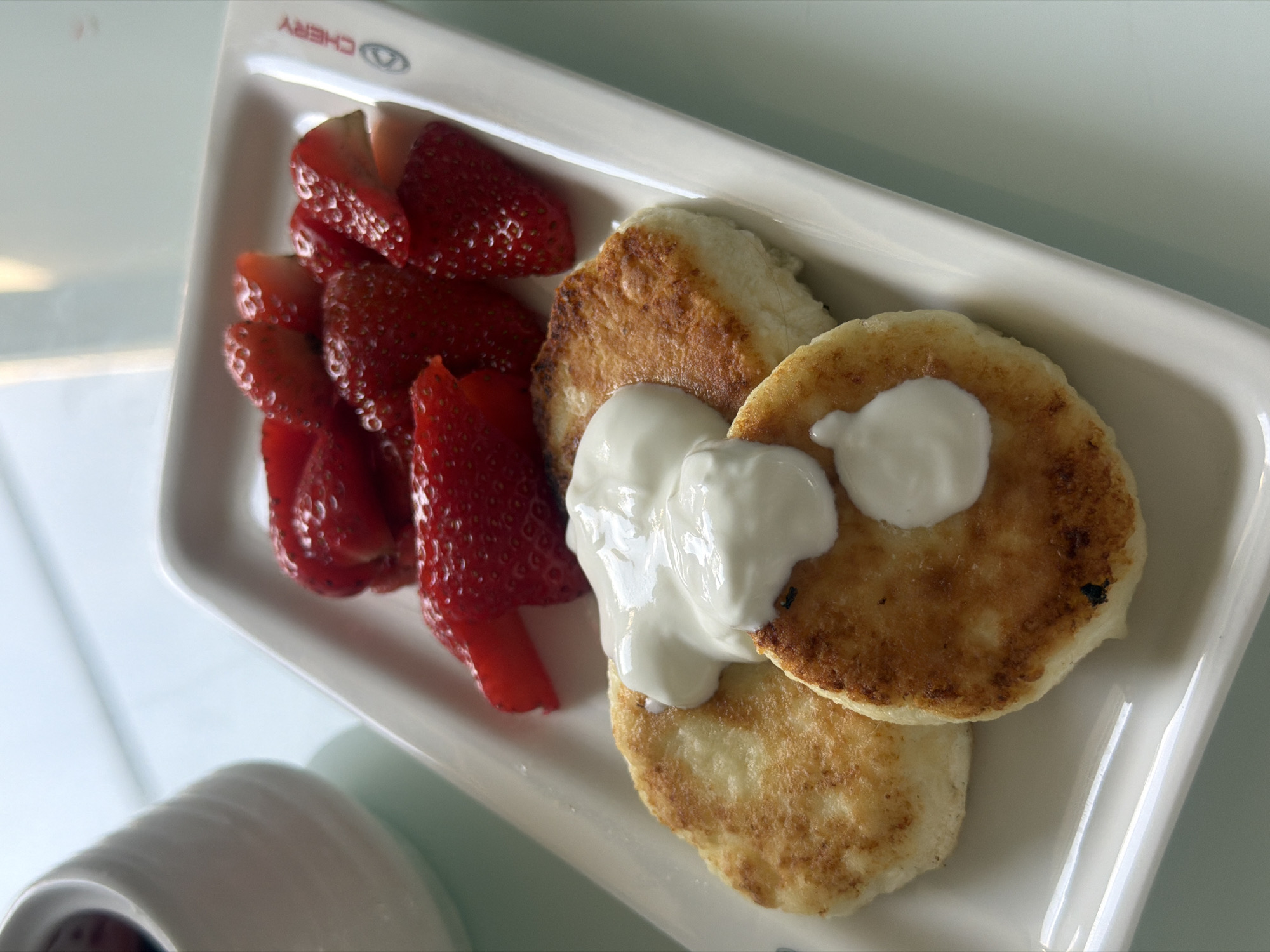 A rectangular white plate displays a breakfast meal. On the left side, a pile of sliced strawberries is arranged. Adjacent to the strawberries are three golden-brown pancakes, topped with a dollop of white sour cream. The plate bears the logo "CHERRY" near the strawberries. The image was likely taken in Rostov-on-Don, Russia.