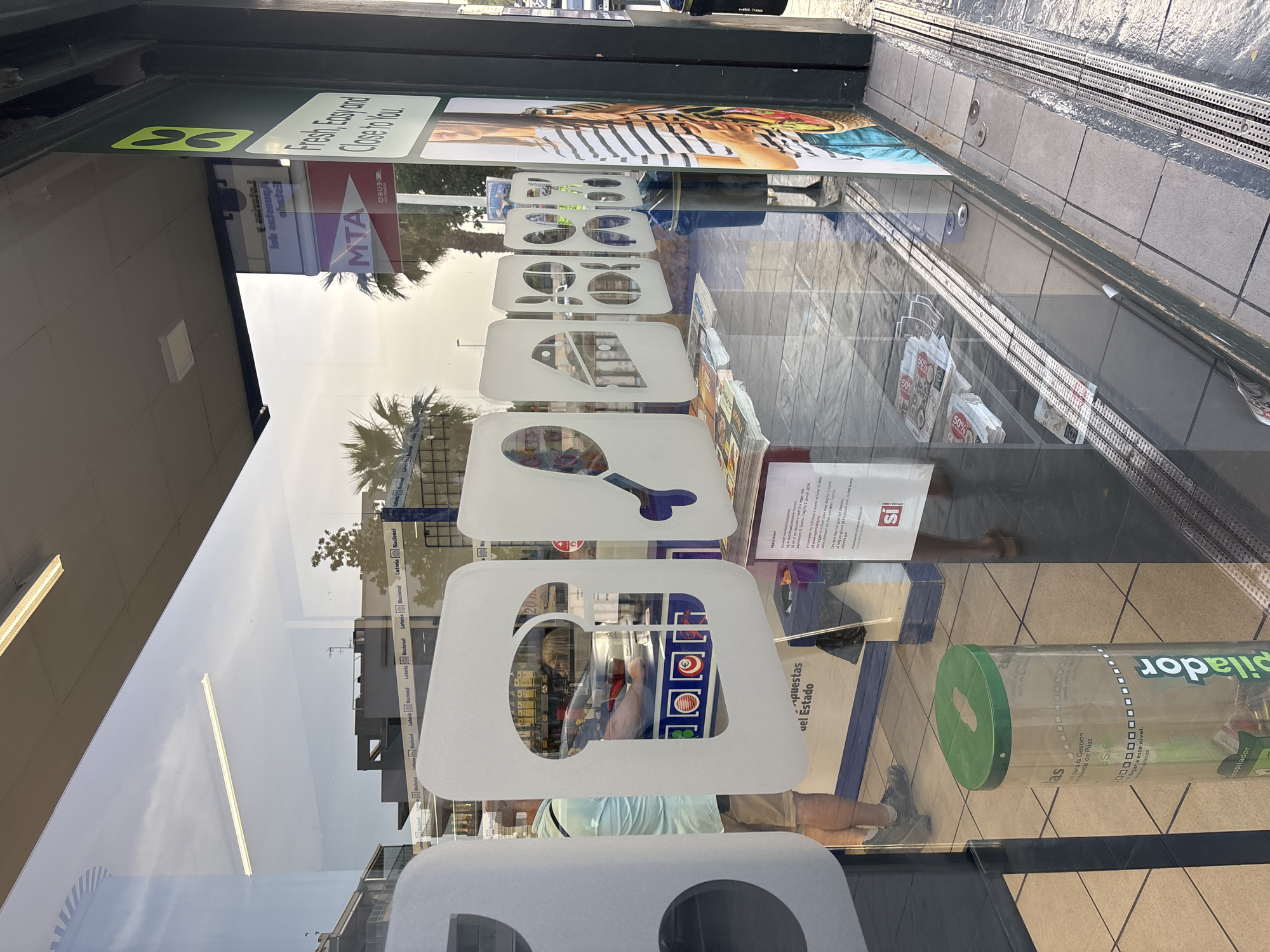 The image shows a street-level view of a shopfront with large glass windows, reflecting the sky and surrounding buildings. The shop appears to be a convenience store or a similar establishment, with various promotional graphics and signage visible both on the interior and exterior.
Inside the shop, several large white cutouts are adhered to the glass, each featuring a distinct icon: a loaf of bread, a chicken leg, and other abstract shapes. Behind these cutouts, shelves stocked with products are visible, along with a person wearing a light blue shirt and beige shorts. A large bin with a green lid and the text "pilador" is positioned near the entrance.
On the right side of the shopfront, a green sign with a yellow leaf-like logo and the text "Fresh, Easy and Close to You." is prominently displayed. Next to it, a poster features a woman with striped clothing.
Reflections on the glass reveal the sky, palm trees, and buildings, suggesting a Mediterranean or similar climate. A red sign with "MTA" is visible in the reflection, indicating a transportation or service provider.
The ground outside the shop is paved with grey tiles and features metal drainage grates along the edge. The overall scene suggests a typical urban commercial setting. The presence of "Urbanización Villa Martín, Spain" indicates the specific location.