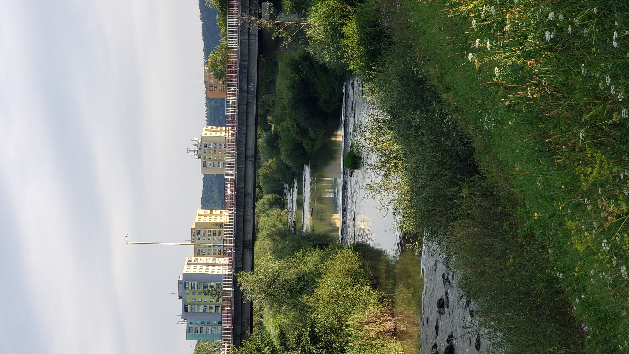 This image depicts a scene in the city of Prešov, Slovakia, featuring a bridge crossing over a river, with apartment buildings visible in the background. The bridge, constructed from concrete, has a white railing along its edge. Beneath the bridge, the river flows calmly, with several small rock formations and a series of man-made cascades or weirs visible in the water. The riverbanks are densely covered with lush green vegetation, including trees, bushes, and tall grasses. In the background, several multi-story apartment buildings, characterized by their colorful facades (yellow, green, and pale yellow), stand against a backdrop of a tree-covered hillside. A solitary lamppost stands tall on the left side of the bridge. The sky above is a light, overcast grey, and the overall lighting suggests it is daytime, likely late afternoon given the quality of light and shadows. No people are visible in the image.