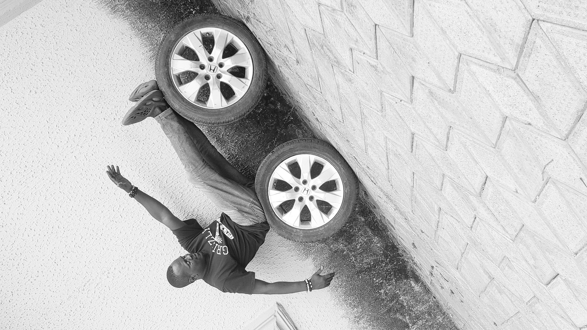 A monochrome photograph captures a man lying horizontally on a textured ground surface, next to a patterned wall, in what appears to be an artistic pose. The man, who is African, wears a dark t-shirt with the text "GRIZZLI" prominently displayed, blue jeans, and unique, textured sandals. He has short hair and bracelets on both wrists. His left arm is extended outwards, palm open, while his right arm is bent, resting near his head. His gaze is directed away from the camera.
Positioned vertically along the patterned wall are two car tires, complete with silver alloy rims. Each rim clearly features a Honda "H" logo in its center, indicating the brand. The ground beneath the man and tires is a rough, uneven texture, possibly asphalt or concrete, with some darker, possibly damp or dirty, patches. The wall is constructed from light-colored blocks arranged in a distinct zigzag or chevron pattern, suggesting a decorative or structural element like a retaining wall or building facade. The image's perspective is rotated, making the wall appear vertical and the man horizontal, lying alongside the tires. The lighting suggests it is daytime. The scene is set in an outdoor, likely urban or semi-urban environment in Sabon Tasha, Nigeria.