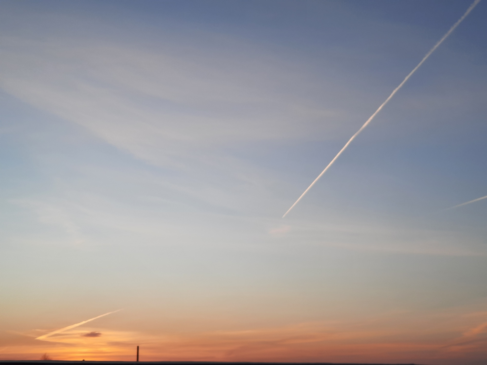 The image shows a sunset sky with several contrails. The sky transitions from blue at the top to orange near the horizon. Three distinct contrails are visible: a long, straight one angling from the upper right, a shorter, fragmented one to the right, and a third near the horizon with a curved end. A dark line suggests the horizon, with a few indistinct vertical features that may be structures or poles. The location is Krasnogorskoye, Russia.