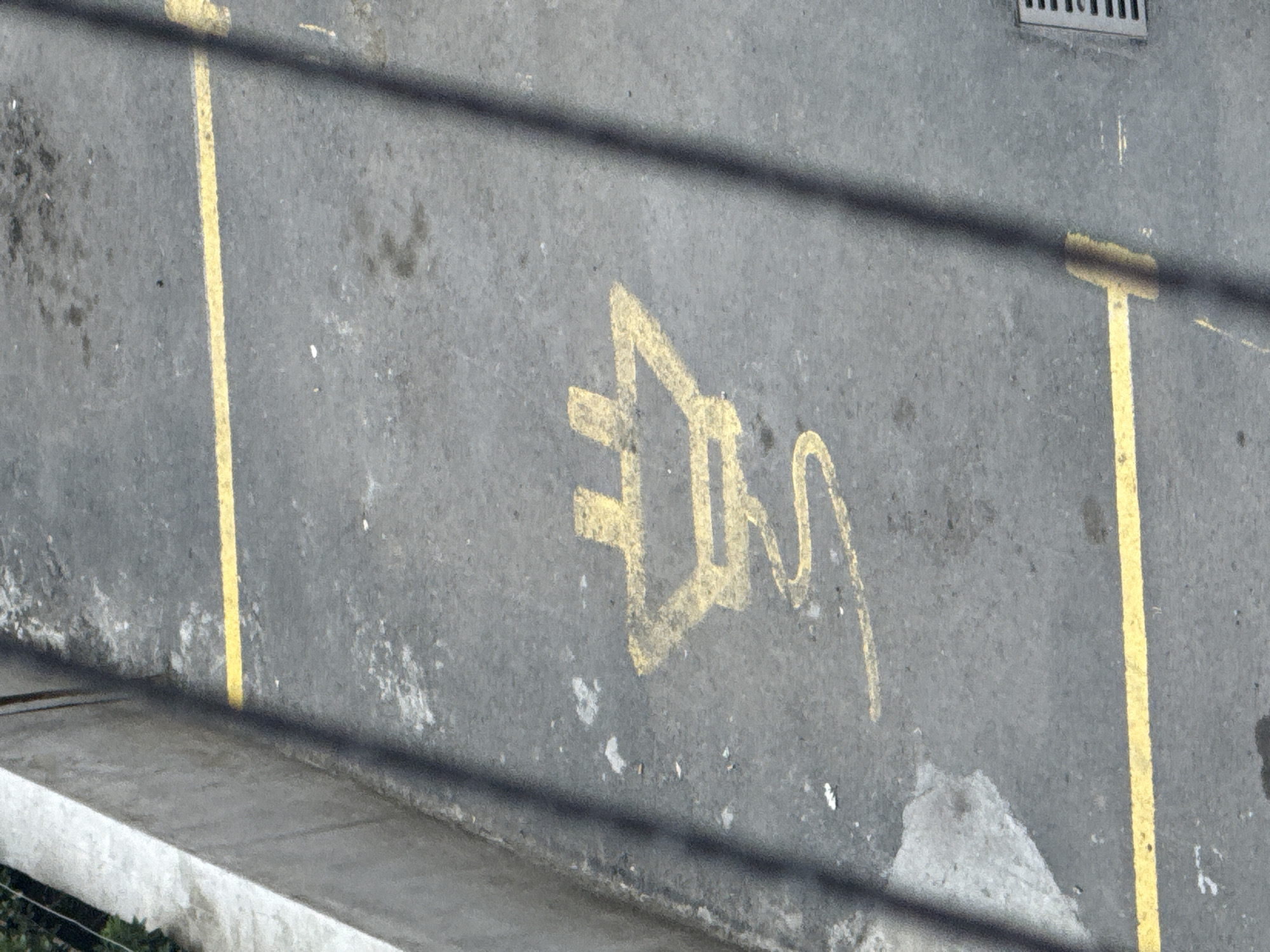 The image shows a parking spot marked with a yellow symbol of an electrical plug on asphalt. The parking spot is delimited by yellow lines. A grey concrete wall runs along the left side of the image. The scene is outdoors, likely in a city, with shadows of wires or branches crossing the image. A drain grate is visible in the lower right corner. The image appears to be taken from an elevated perspective. The location of Tbilisi, Georgia is not explicitly indicated in the image but is inferred from the prompt.