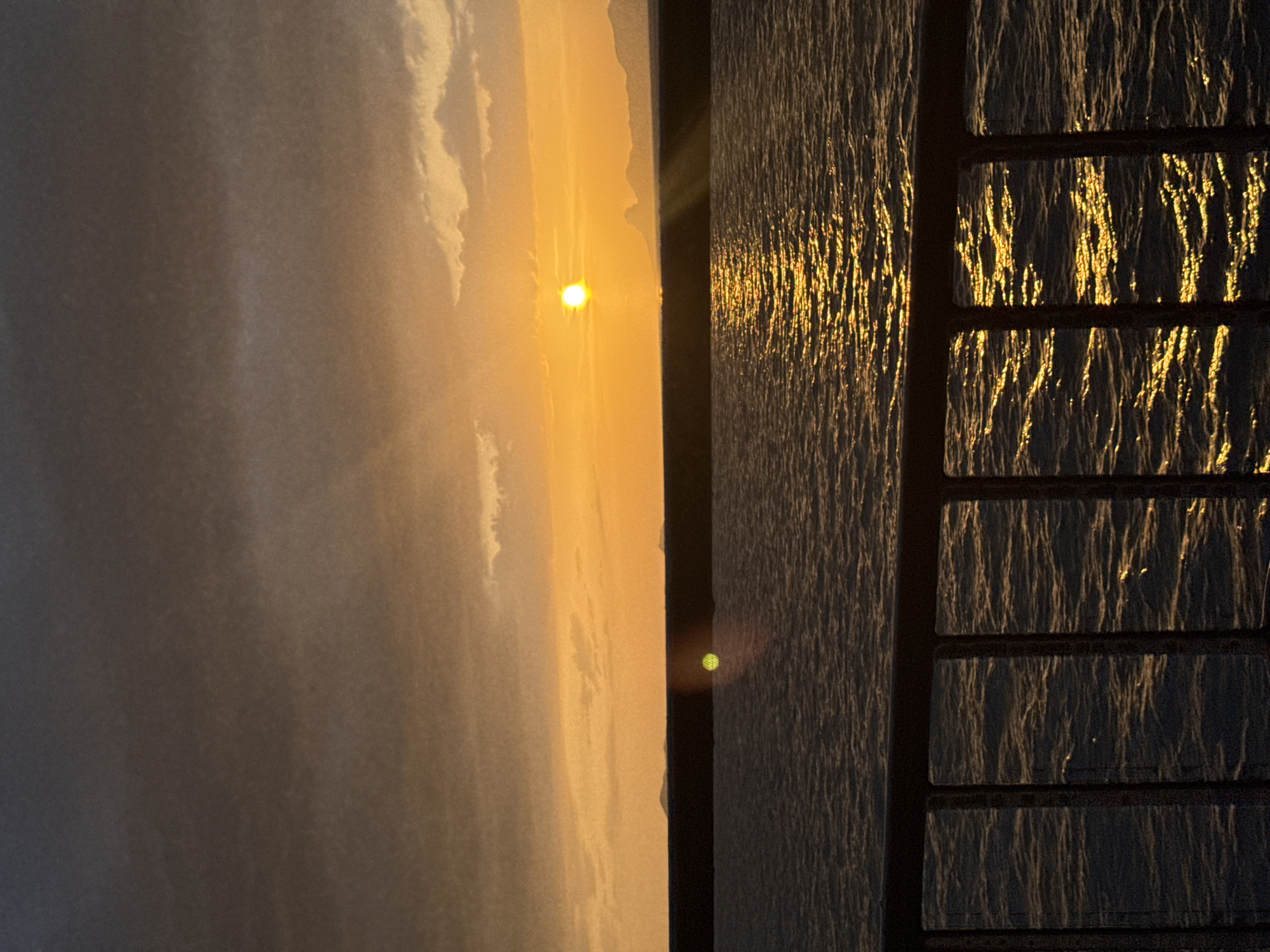 The image depicts a sunset over the ocean, viewed from behind a metal railing. The sun, a bright yellow orb, is partially obscured by the horizon, casting a golden light across the water. The sky is a gradient of orange and gray, with clouds scattered across it. In the distance, the silhouettes of islands or mountains can be seen. The water is choppy, with reflections of the sun creating streaks of light on its surface. The railing in the foreground is dark and appears to be made of metal. The overall mood of the image is tranquil and serene.