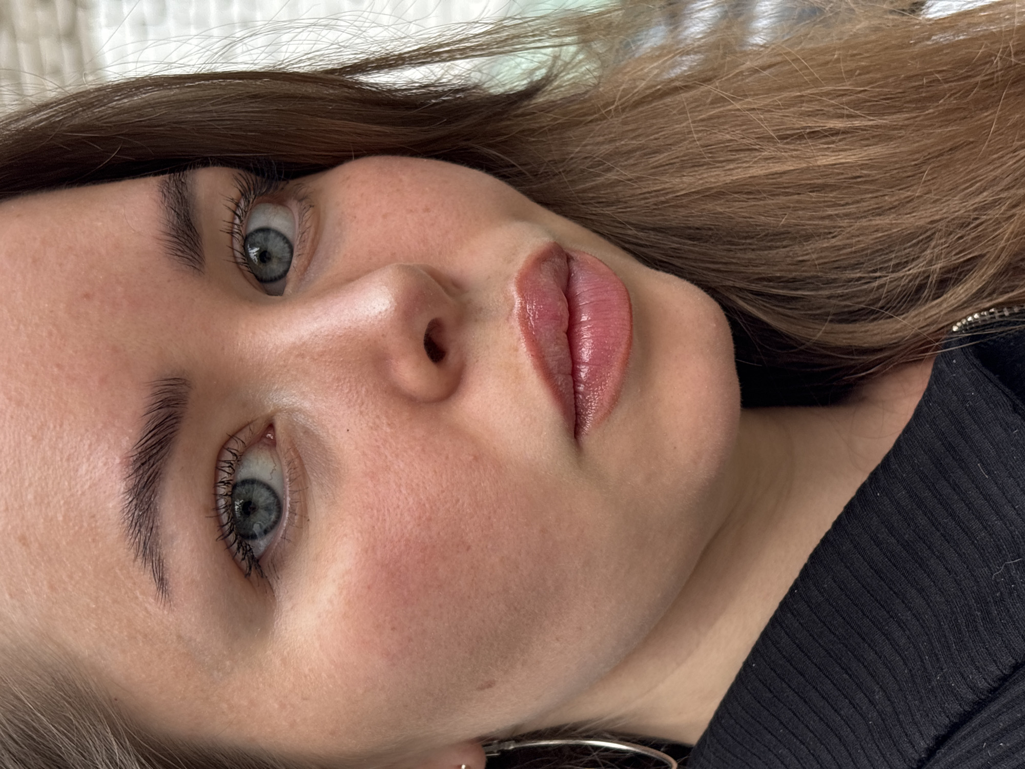 A close-up, slightly angled shot of a woman's face. She has fair skin, light brown hair, and grey-blue eyes with mascara. Her lips are full and have a pinkish-red color, possibly from lipstick or lip tint. She is wearing a black shirt. The background is slightly blurred. There's no indication that the photograph was taken in Izhevsk, Russia.