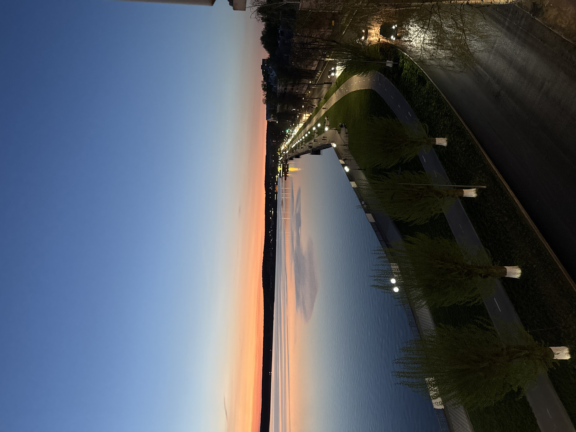 The image shows a nighttime or twilight view of a riverfront area in Votkinsk, Russia. The sky transitions from blue to orange near the horizon. A body of water reflects the colors of the sky. Along the riverbank, there is a walkway illuminated by evenly spaced lights. Trees line the walkway. A road runs parallel to the walkway and is visible on the right. The lighting suggests that it is dusk or early evening. No people are clearly visible.