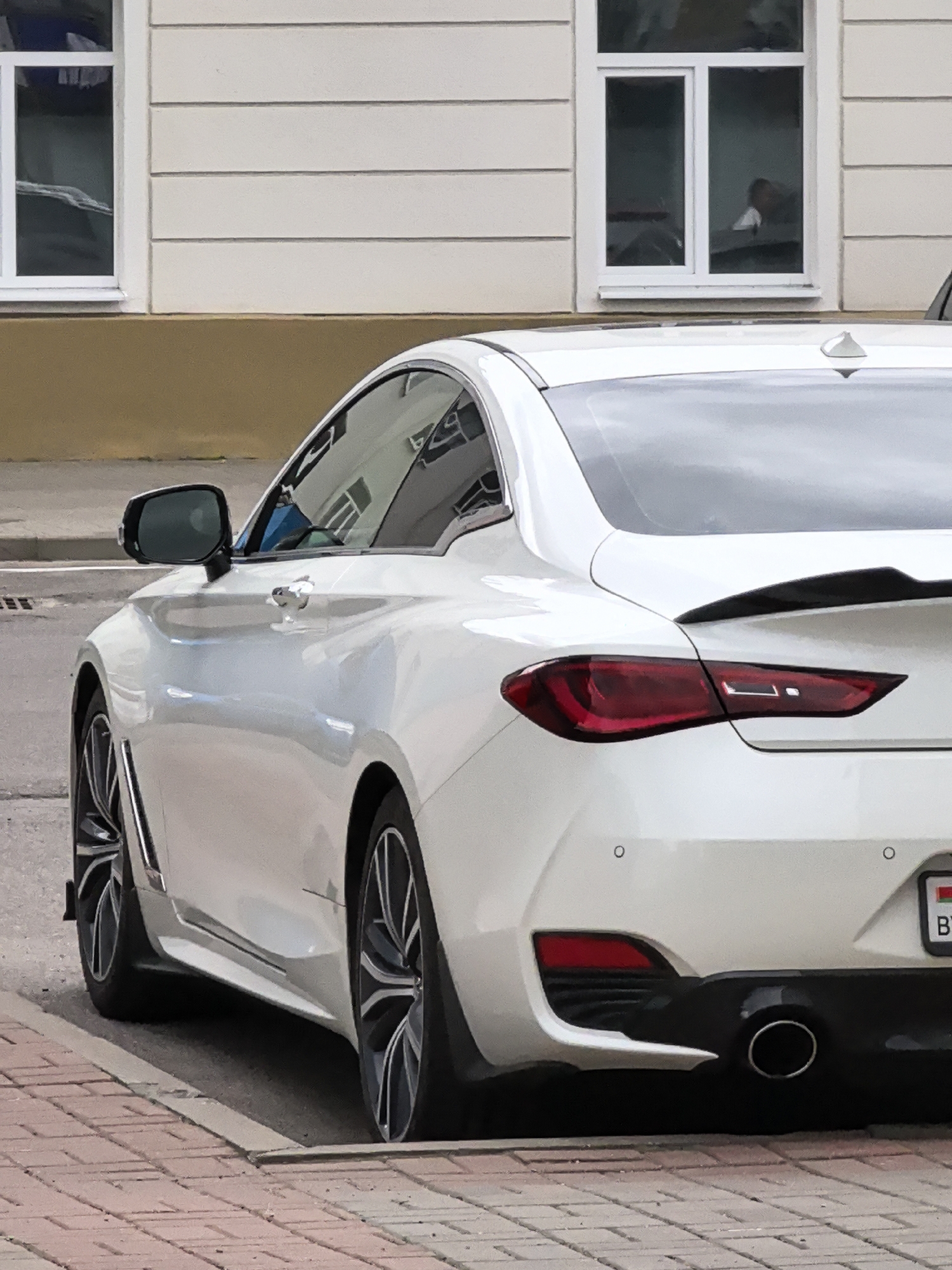 A white Infiniti Q60 is parked on a brick sidewalk next to a street. The car is viewed from the rear passenger side, showing its sleek design and tinted windows. The background features a building with beige siding and white trim, along with two large windows. One window reflects an interior with a person visible. A license plate with the letters "BY" and a Belarus flag is attached to the rear of the car. The car's wheels have a multi-spoke design.