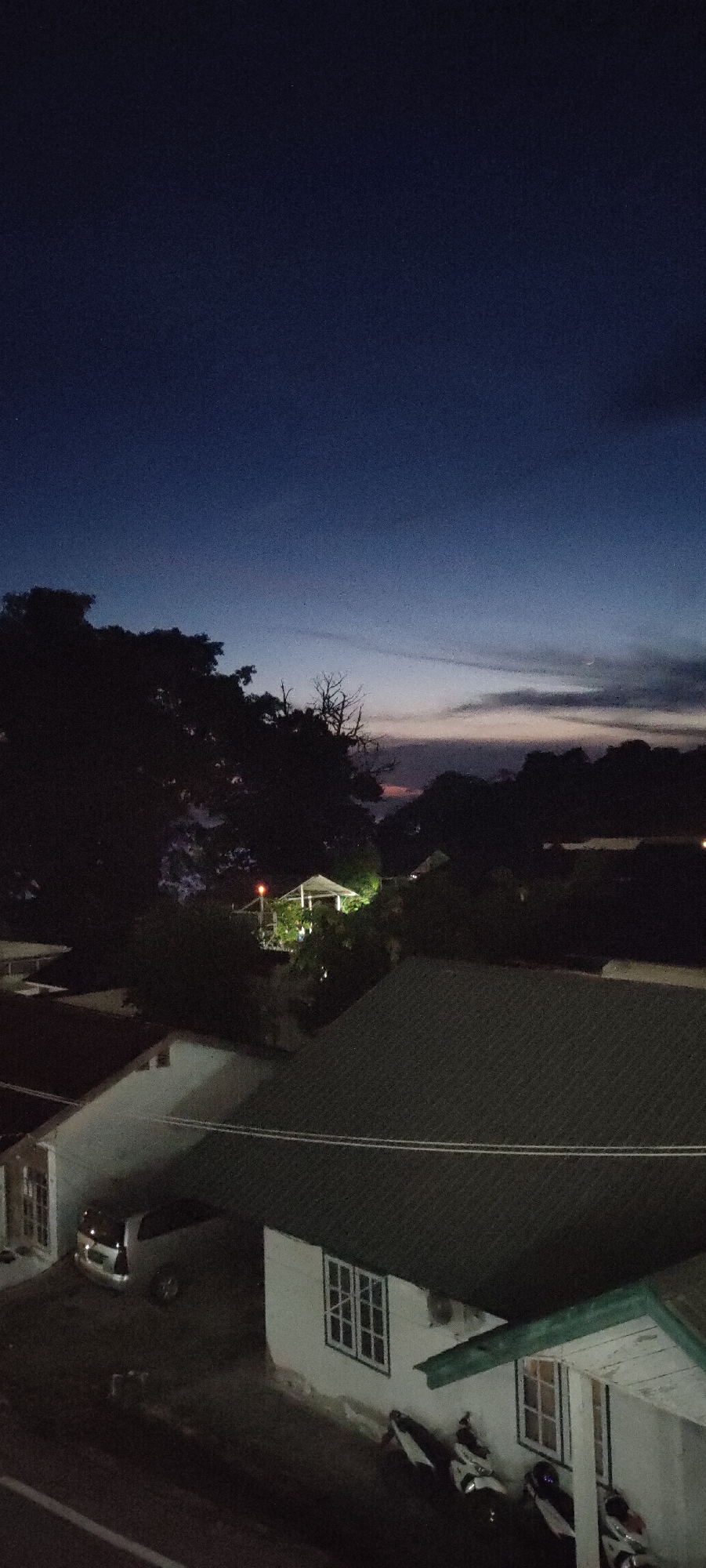 A low-angle, vertical shot captures a dusky sky over a residential area in Sabang, Indonesia. The sky transitions from a deep navy blue at the top to lighter shades of blue and pink near the horizon, with faint clouds visible. Silhouetted against the sky are dense trees on the left and a sparser line of trees and foliage across the mid-ground.
In the mid-ground, a small, open-sided structure, possibly a gazebo or pavilion, is illuminated by warm light. A red light is visible on top of this structure. Below the structure and further in the foreground, several buildings with dark rooftops are visible. One building in the lower left is white with visible windows. A silver minivan is parked in front of this building. To the right, another white building with green trim is prominent, featuring multiple windows. Parked in front of this building are at least three motorcycles, two of which are white and black, and another is partially visible. Power lines are strung across the foreground. The overall scene suggests an evening setting in a somewhat rural or suburban part of Sabang.