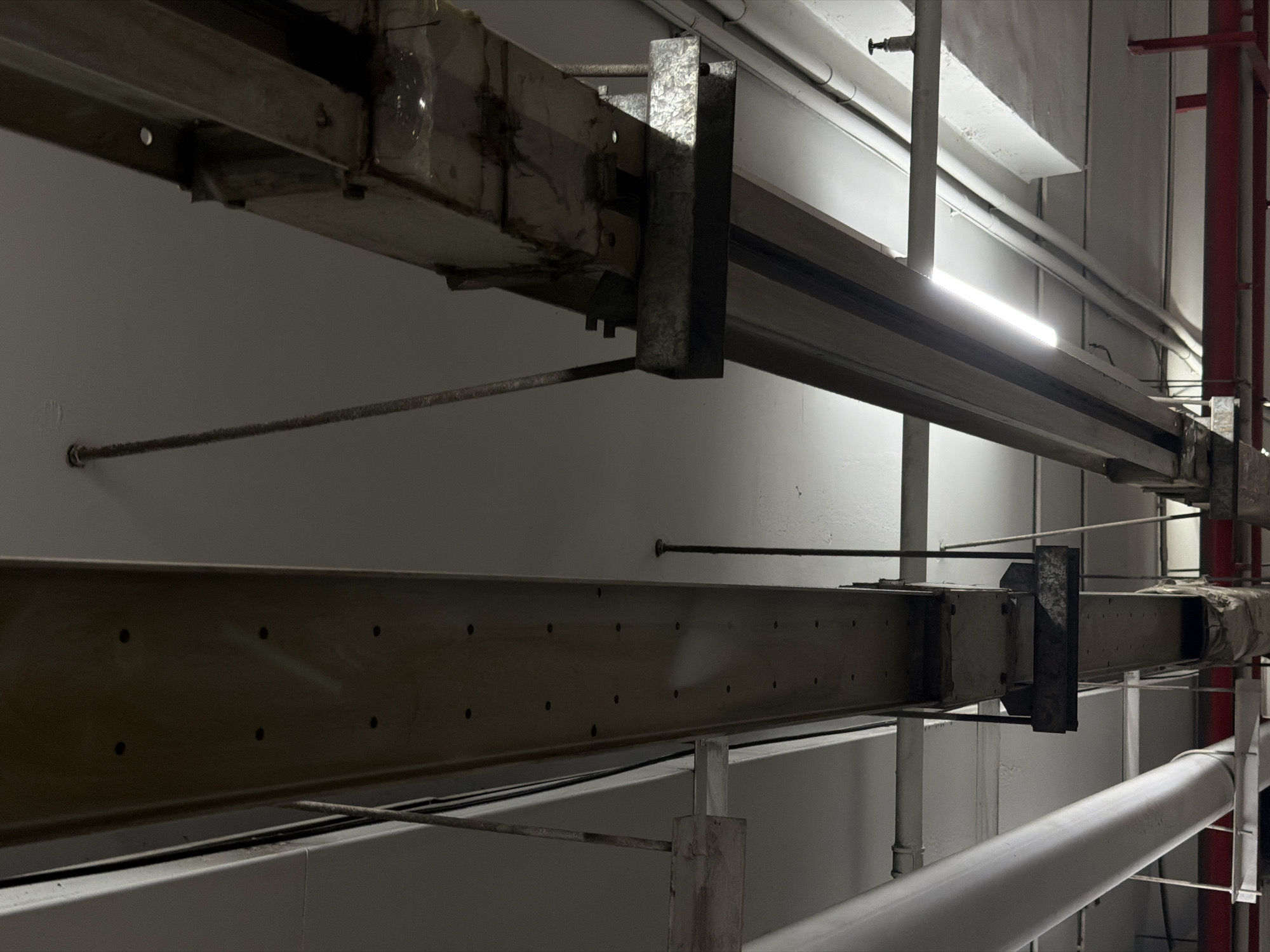 An interior shot, likely taken in a dimly lit industrial or commercial space in Da Nang, Vietnam, focuses on a ceiling with exposed infrastructure. Two prominent, heavy beams run horizontally across the frame, suspended from the ceiling by metal rods and supports. The beam on the left appears to be a large, rectangular conduit or channel, its surface marked with numerous small circular indentations. It is suspended by thin wires or cables. The beam on the right is more complex, a multi-layered structure with visible metal components and what appears to be insulated wrapping on its underside. This beam is also suspended by metal rods.
Beneath and between these main beams, various pipes and conduits are visible. On the left, a large, white cylindrical pipe runs horizontally, supported by metal brackets. Other smaller pipes, possibly electrical conduits or ventilation systems, are also present, some running parallel to the main beams and others intersecting them. A bright, linear fluorescent light fixture is mounted beneath the right-hand beam, illuminating a portion of the area with a stark white glow. In the background, sections of a white ceiling are visible, along with a hint of red structural elements, possibly shelving or support beams, at the bottom of the frame. The overall impression is one of functional, utilitarian construction, typical of infrastructure in service or utility areas. There are no people visible in the image.