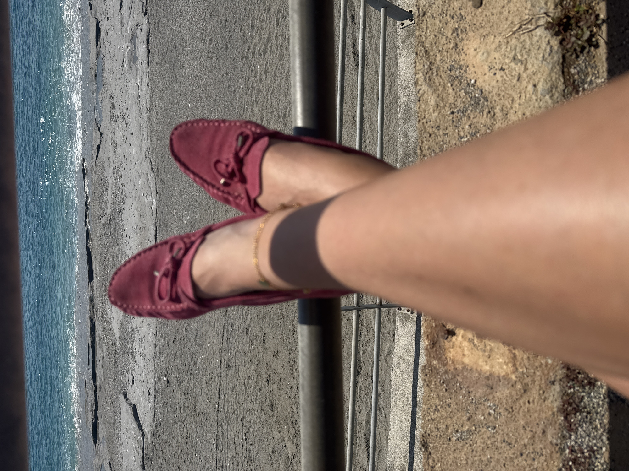 The image shows a first-person perspective of a person sitting with their legs crossed over a metal railing, overlooking a beach. The person is wearing maroon suede loafers and a gold ankle bracelet. The beach has dark sand, and the ocean is visible in the background. The railing is mounted on a concrete ledge with a rough surface. The location is Unknown, Unknown.