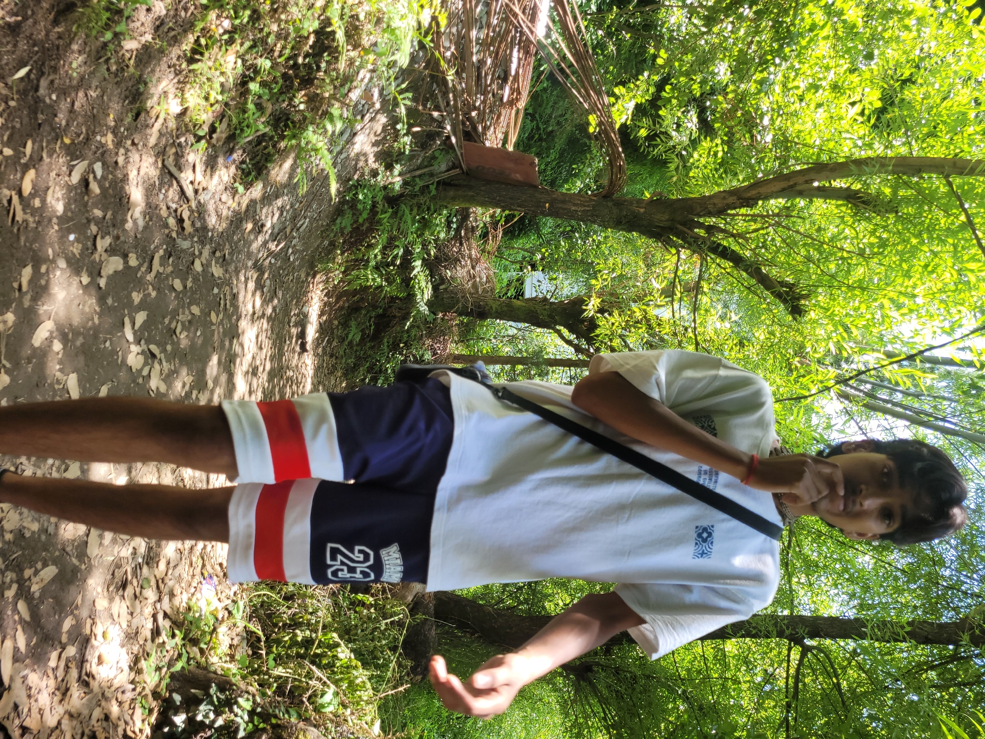 A young man stands on a dirt path in a wooded area, likely Vashisht, India. He is wearing a white t-shirt with a blue graphic and a black strap across his chest, along with blue shorts that have red and white stripes at the hem and the number "23" on the left leg. His right hand is near his face, and his left arm is bent, with his hand extended. The path is covered with fallen leaves and debris, and sunlight filters through the dense green foliage of trees that surround the area. To the left of the path, a pile of rusted metal rods and some wooden planks are visible against a tree trunk. The overall scene suggests a natural, outdoor setting with elements of what appears to be construction materials nearby.