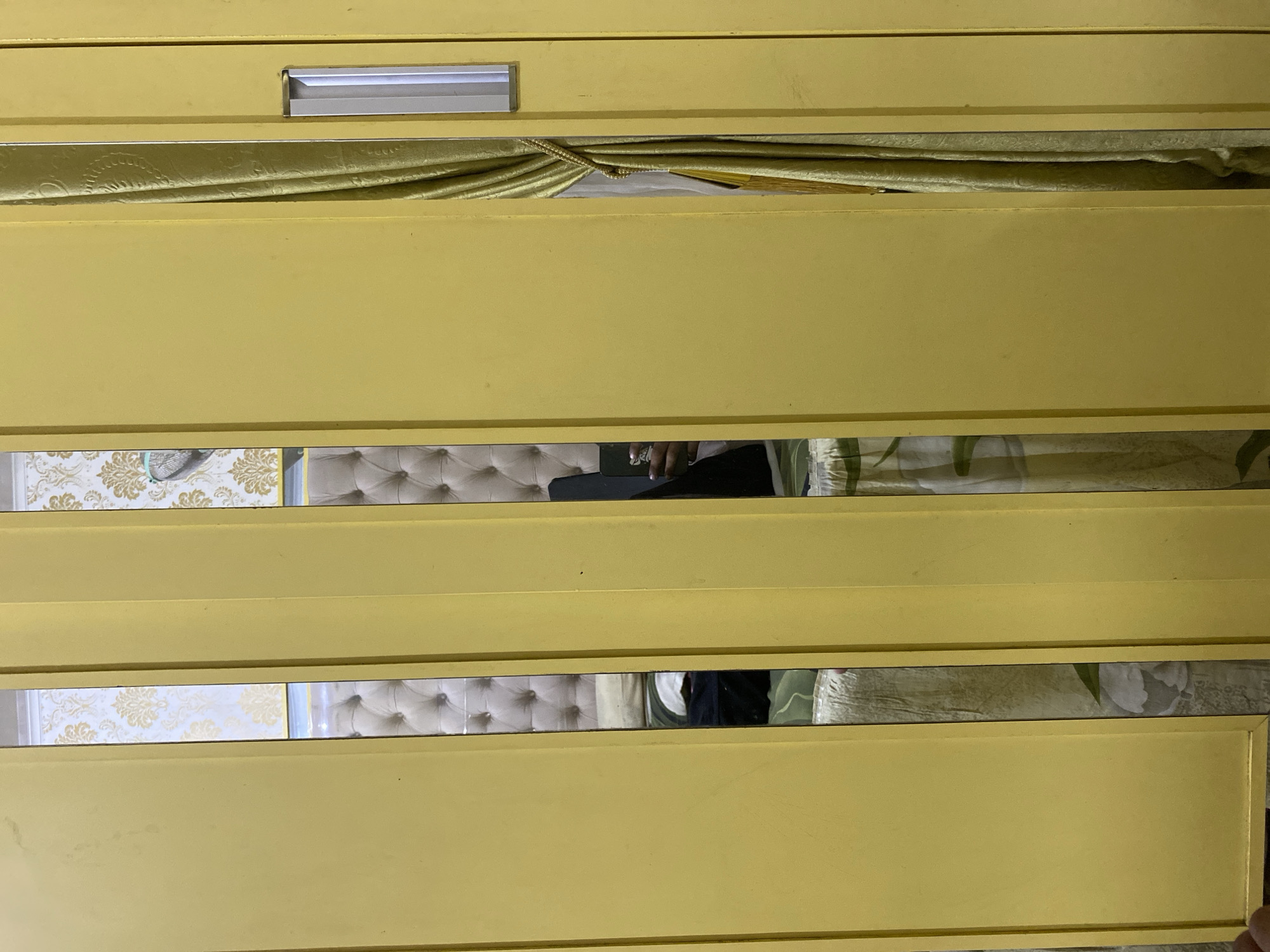 The image shows a section of a yellow wardrobe with mirrored doors. The mirrors reflect a bedroom interior, including a tufted headboard of a bed and patterned wallpaper. In the rightmost mirror, a person's arm and hand holding a phone are visible. A gold-colored curtain is also visible in the reflection of the right side of the wardrobe. The wardrobe appears to be a sliding door type, with vertical divisions between the mirrored panels. The scene is indoors, likely a bedroom in a residential setting.