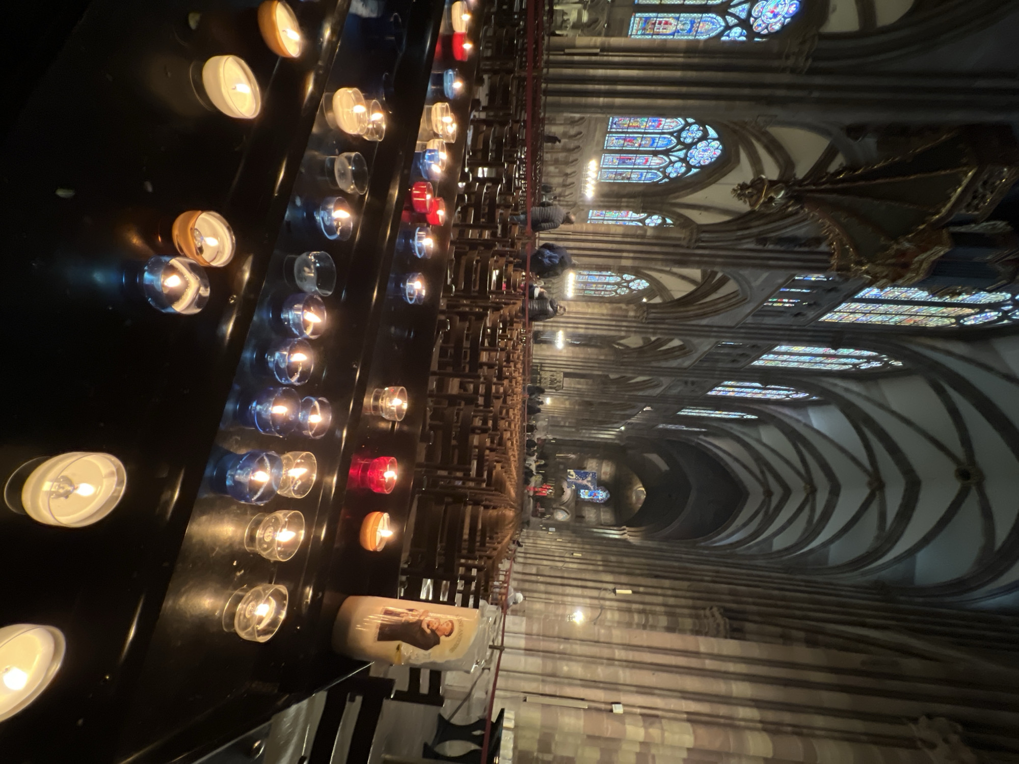 The image shows the interior of Strasbourg Cathedral in Strasbourg, France. A line of lit candles in various colored glass holders sits in the foreground. Beyond the candles, rows of empty wooden pews stretch into the distance. Stained glass windows are visible near the ceiling. Several people are seated in the pews. A decorative sculpture is visible near the top right corner. The architecture features arched ceilings and stone columns.