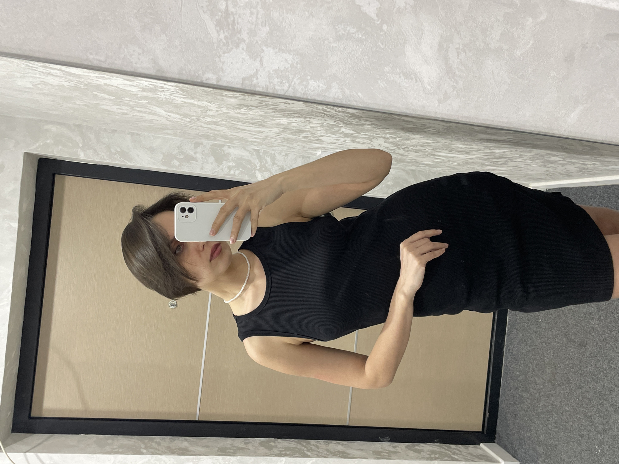A woman is taking a selfie in front of a mirror. She is wearing a black dress and a white pearl necklace. She has short, dark hair. The background is a wall and a door. The location is not identifiable from the image.