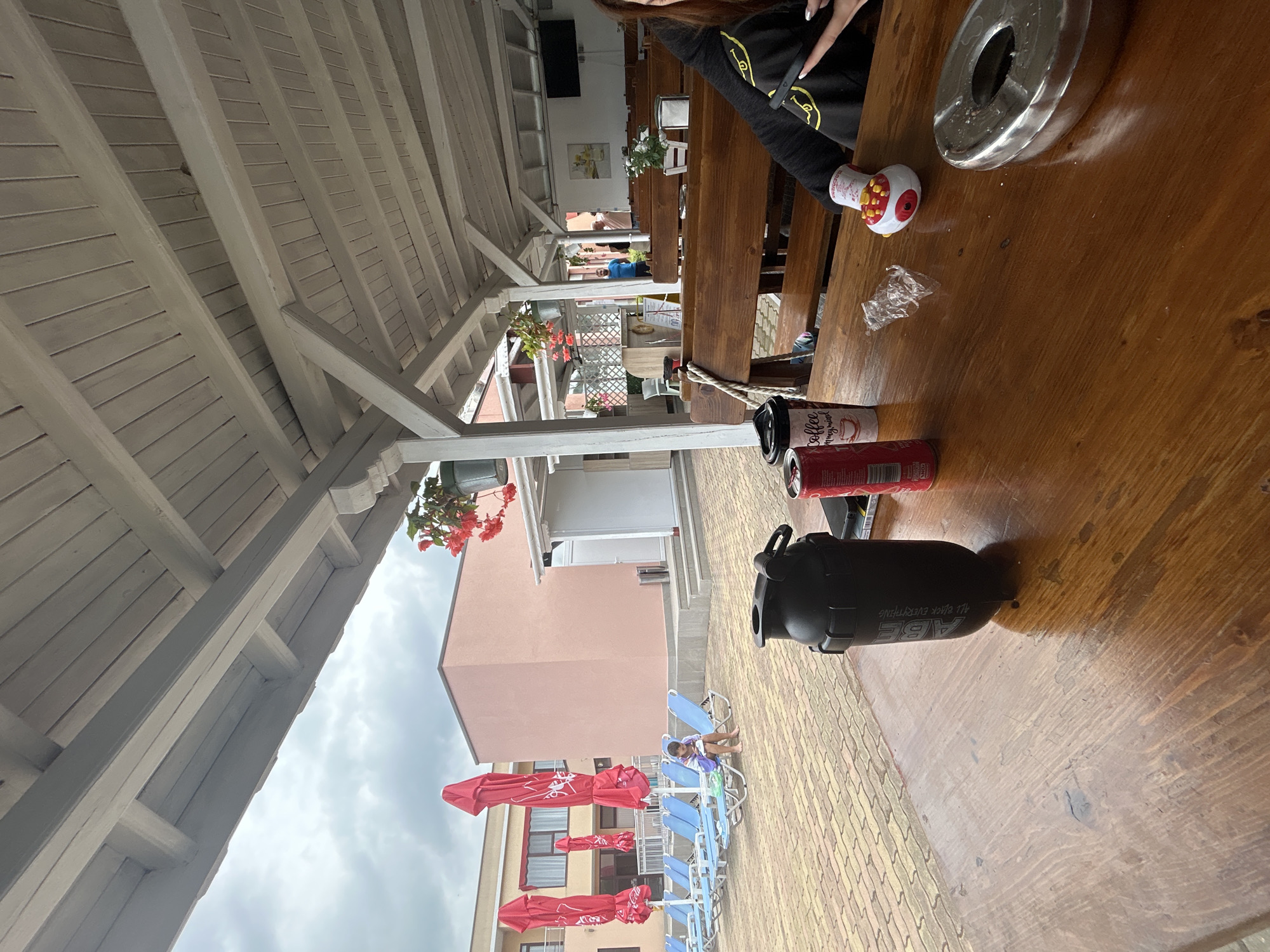 A medium shot captures a rustic wooden table at an outdoor cafe, sheltered by a white-painted wooden canopy. The table is set with a black shaker bottle, a red soda can, a disposable coffee cup, an ashtray, and a small toy. In the background, lounge chairs and red umbrellas are arranged on a patterned brick patio, adjacent to a pink building. A child sits on one of the lounge chairs, facing away from the camera. The sky is overcast.