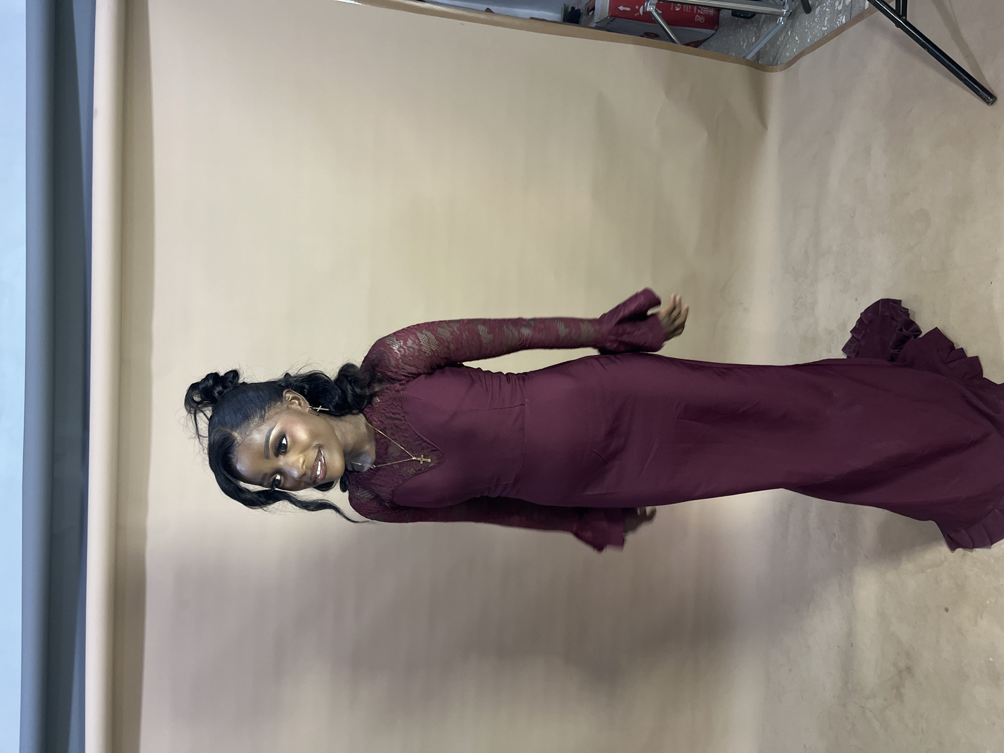 A young woman stands against a beige backdrop, smiling and looking to her left. She is wearing a long, burgundy dress with lace sleeves that flare out at the wrists. The dress has a sweetheart neckline and a floor-length skirt with ruffled trim. She has her hair styled in a high bun with loose tendrils framing her face. A gold cross necklace is visible around her neck. The environment appears to be a photo studio, with studio lighting equipment visible on the right side of the frame. The backdrop is a seamless paper roll, common in photography settings. The woman's makeup is visible, including eyeshadow, eyeliner, and lipstick. The lighting is soft, highlighting her features and the details of her dress. The overall mood is elegant and celebratory.