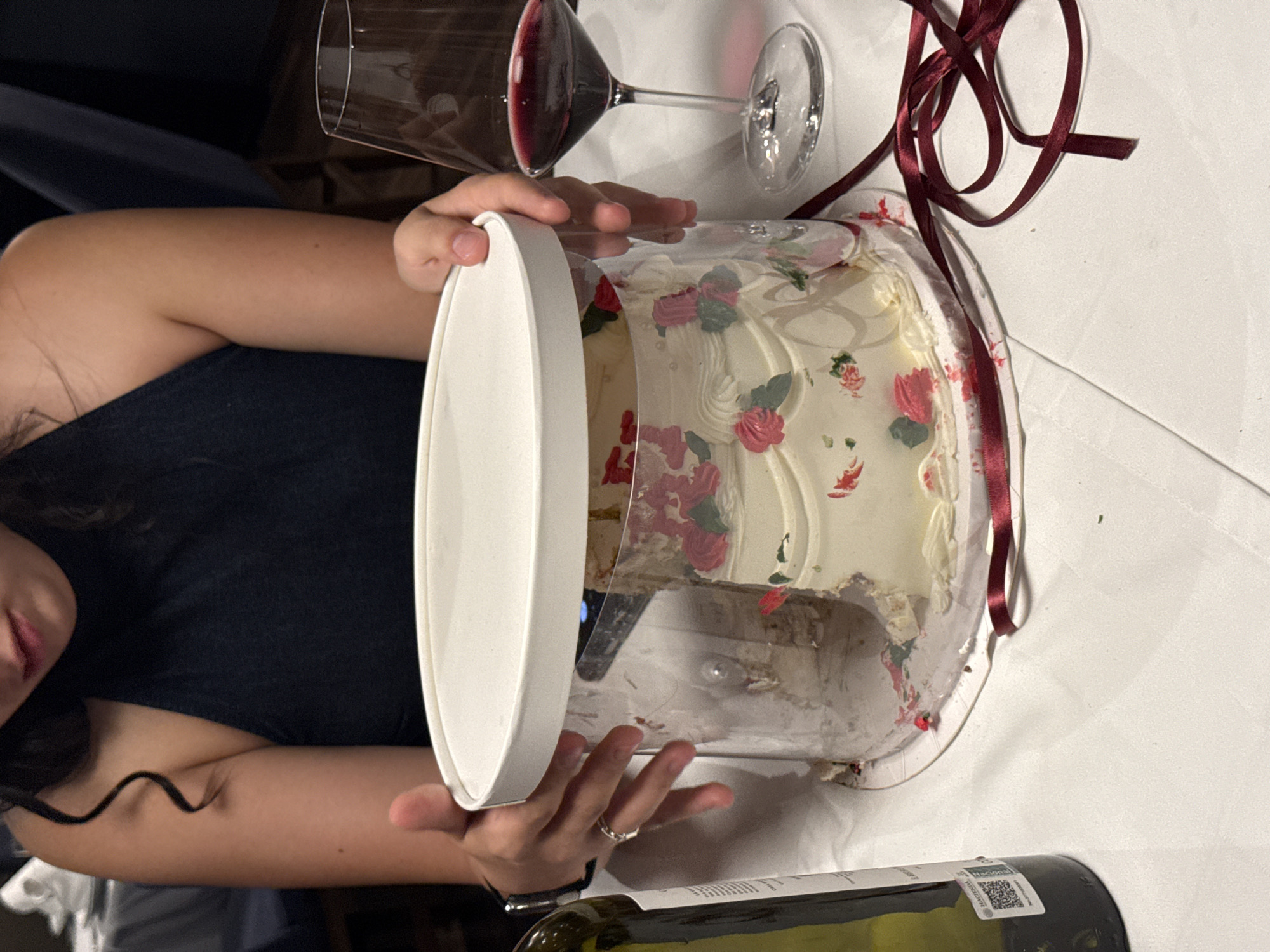 A person is holding a clear plastic cake container with a white lid. Inside is a partially eaten cake with white frosting and red flower decorations. Next to the cake is a glass of red wine and a bottle of wine. A maroon ribbon is draped around the cake container. The scene is set on a white table. The location appears to be an indoor setting, but the specific city of Puebla, Mexico cannot be confirmed from the image.