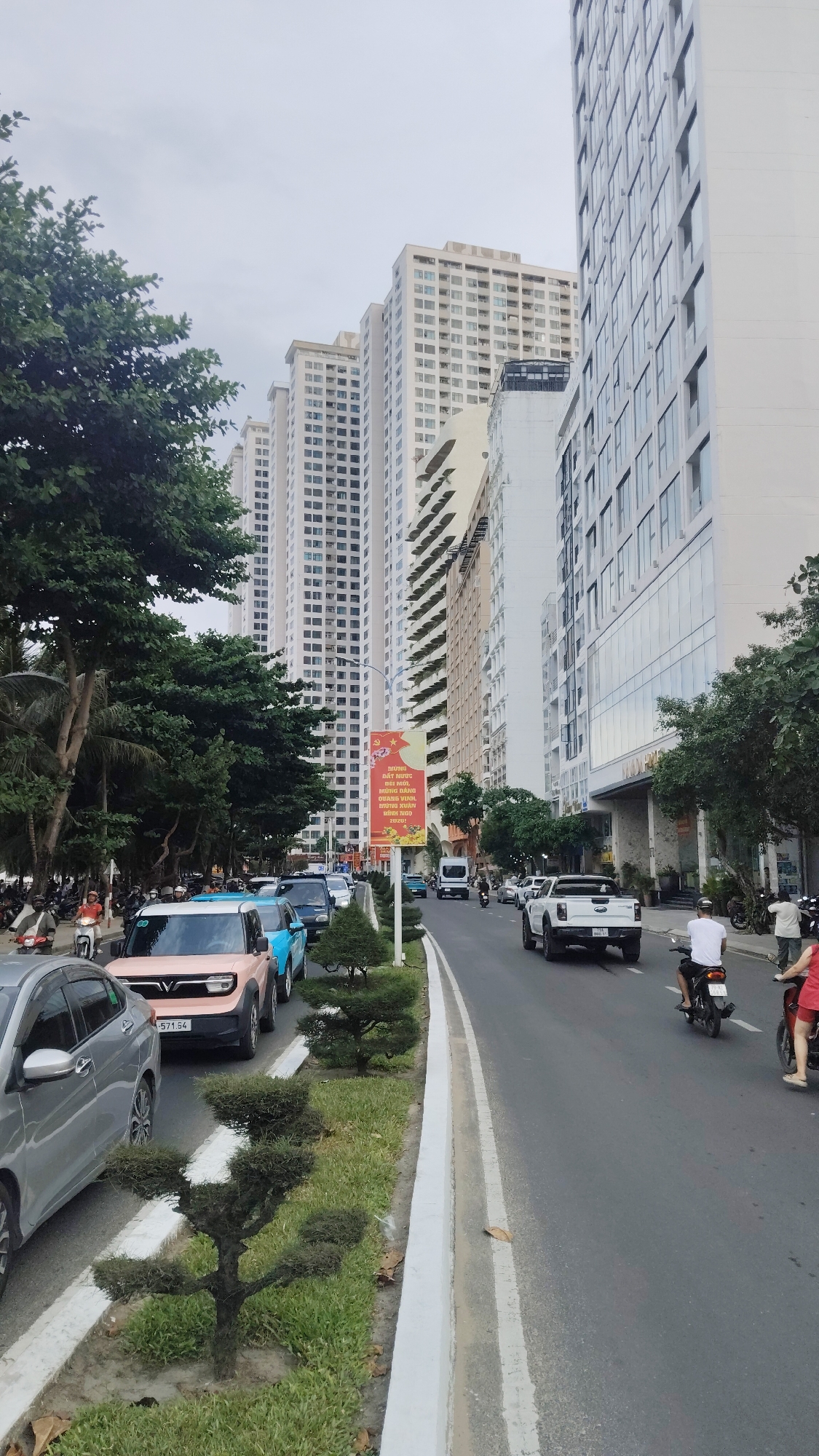The image depicts a street scene in Bac Nha Trang Ward, Vietnam. A row of high-rise buildings lines the right side of the street, with trees and lower buildings to the left. A street divider with small, sculpted trees runs down the center of the road. Several cars and motorcycles are visible in both lanes. The sky is overcast. A banner with Vietnamese text and imagery is placed along the street divider. People are seen walking and riding motorcycles. Vehicle number plates are visible.
