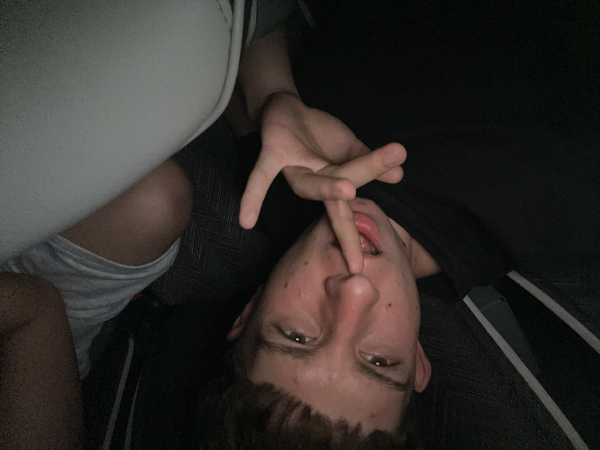A young, fair-skinned male lies upside down, with a finger inserted into his mouth. He is wearing a dark-colored shirt with white stripes and light grey shorts. The background is mostly dark and obscured, with a white cushion or object partially visible. The location of the photo is not discernible from the image, and there is no indication it was taken in Saraia, Bulgaria.