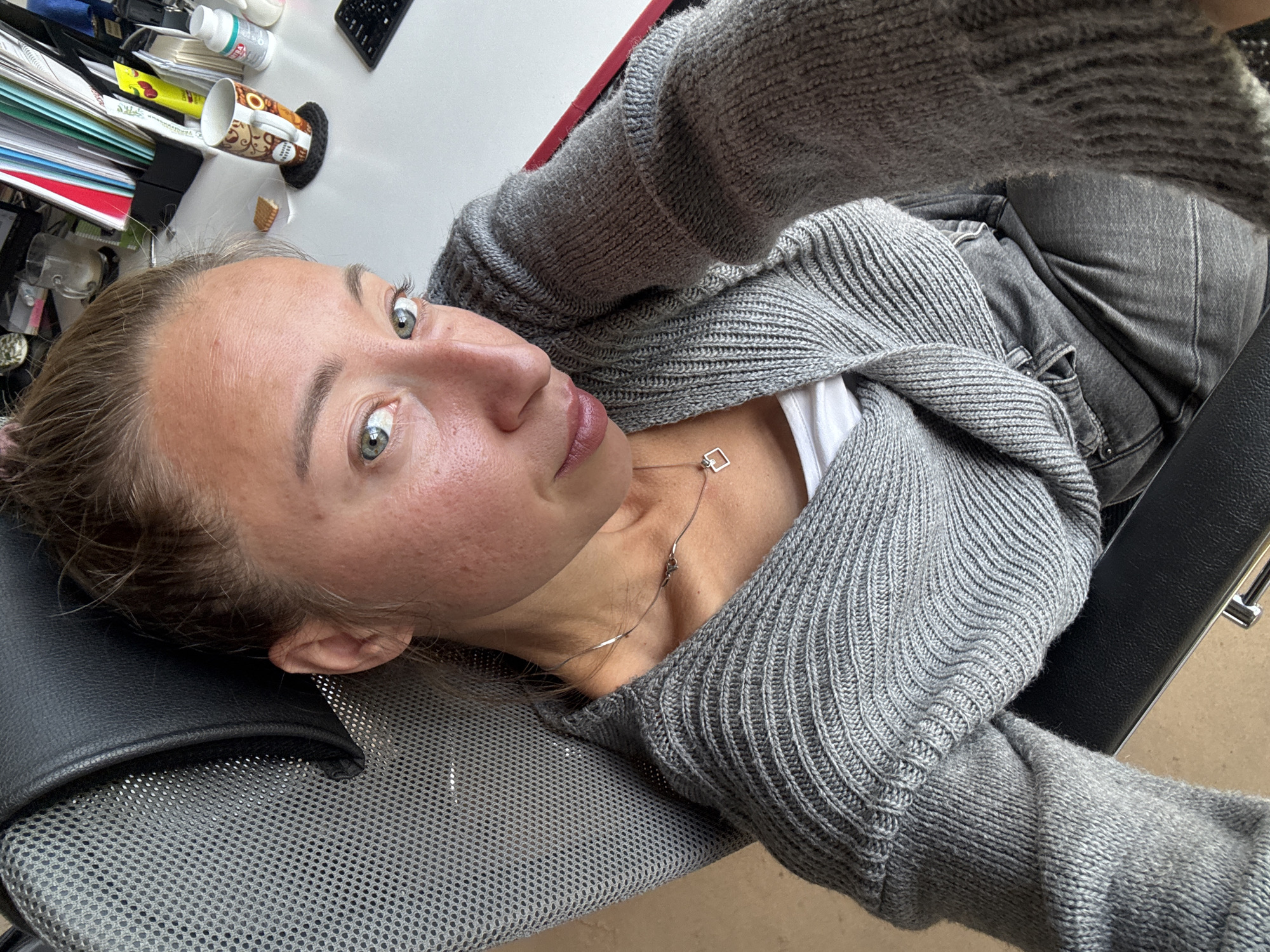 A woman is sitting in a chair in an office. She is wearing a gray sweater and a white shirt. Her hair is pulled back in a bun. She is looking at the camera. There is a desk in the background with a computer, papers, and a mug. The scene appears to be indoors.