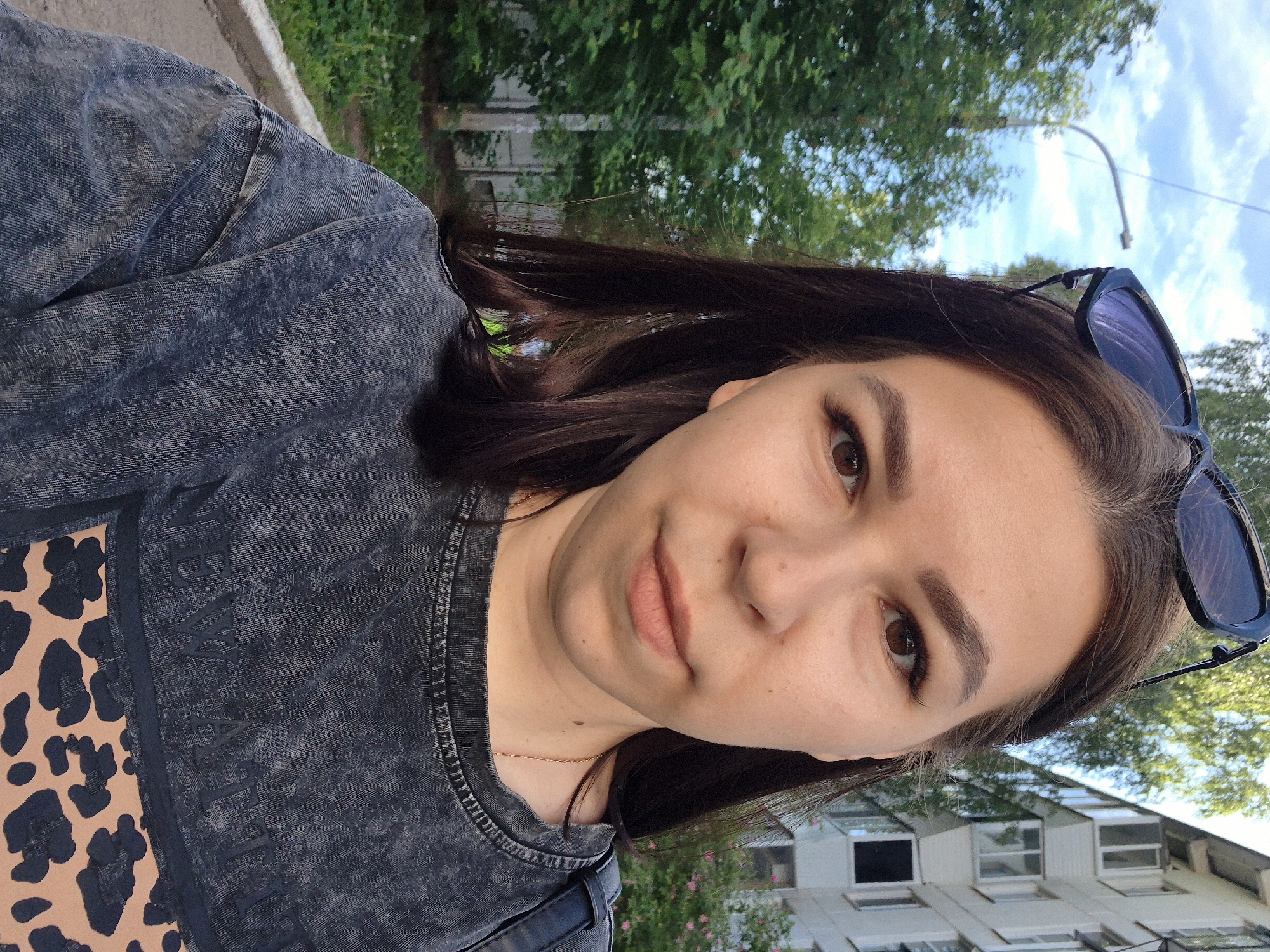 The image displays a young woman taking a selfie outdoors in Chaikovsky, Russia, during the daytime. She has dark brown hair, brown eyes, and a fair complexion, with a neutral expression and a slight smile. Dark sunglasses are perched on her head.
She is wearing a dark gray, acid-wash or stonewashed t-shirt or sweatshirt. The garment features a light brown panel with a dark leopard print design on the left side. Adjacent to this, stylized, textured text is visible on the dark gray fabric, which appears to spell "DMYAT" or a similar word. A thin gold chain necklace is visible around her neck.
The background shows a clear blue sky with some white clouds and lush green trees, indicating pleasant weather. To her right, a multi-story building with numerous windows is visible, suggesting an urban or residential setting. A sidewalk or paved area with a curb can be seen on the far left of the frame.