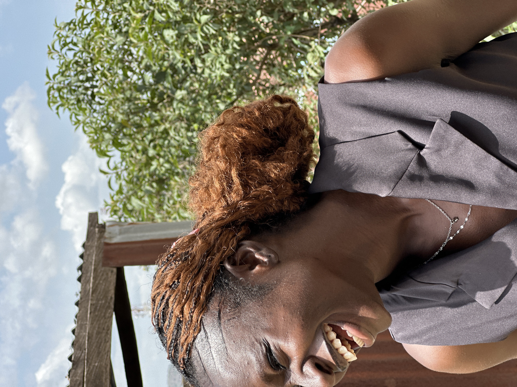 A close-up, profile shot captures a Black woman with braided, auburn-tinted hair pulled into a bun, laughing outdoors in Jos, Nigeria. She is wearing a dark grey sleeveless blazer over a silver necklace. The background shows a corrugated metal roof structure, lush green foliage, and a cloudy blue sky. The lighting suggests it is daytime.