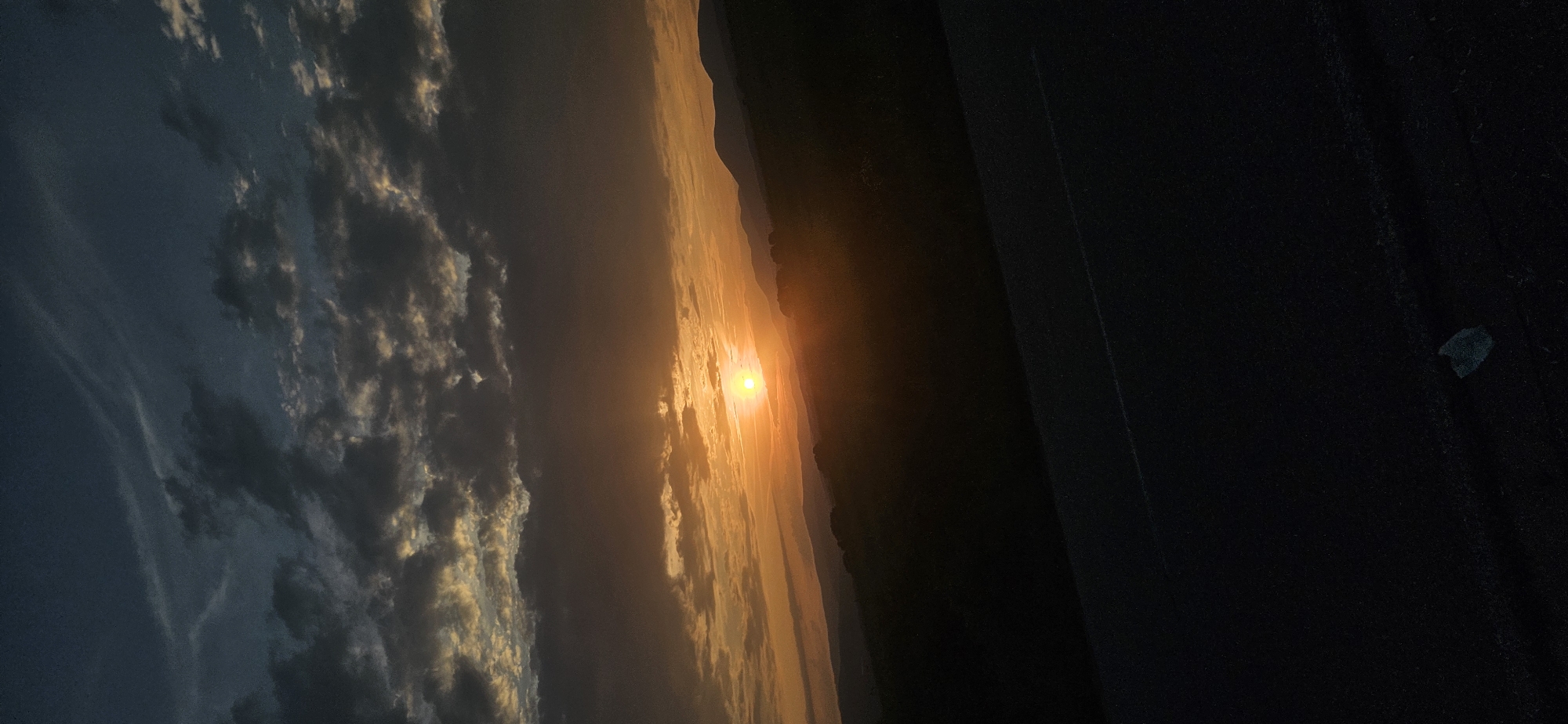 This vertical image depicts a sunset over a mountainous landscape, observed from what appears to be a road or elevated surface in Momin Prohod, Bulgaria. The sky dominates the upper two-thirds of the frame, transitioning from a deep twilight blue at the very top to vibrant oranges and yellows near the horizon where the sun is positioned. Numerous clouds are present; those higher up are dark grey and blue, while lower clouds are illuminated from beneath by the setting sun, displaying warm orange and gold hues.
The sun, a brilliant orange-yellow sphere, is visible on the horizon, slightly left of the center, casting a strong glow that diffuses through atmospheric haze. Below the horizon line, multiple layers of hills and mountains stretch across the midground. These landforms appear as progressively darker silhouettes, with the most distant peaks faintly outlined against the illuminated sky and the closer hills appearing as dark, almost black masses due to the low light and shadows. Sparse vegetation is discernible on some of these closer hills.
The foreground, occupying the bottom third of the image, consists of a dark, textured surface, likely asphalt, suggesting a road or viewing point. A faint, thin white line runs horizontally across this surface, indicative of a road marking. In the bottom right corner, a small, irregularly shaped piece of dark debris with a subtle blue tint is visible on the ground. No people, animals, or active subjects are present in the scene. The overall lighting suggests the crepuscular hours of dusk.