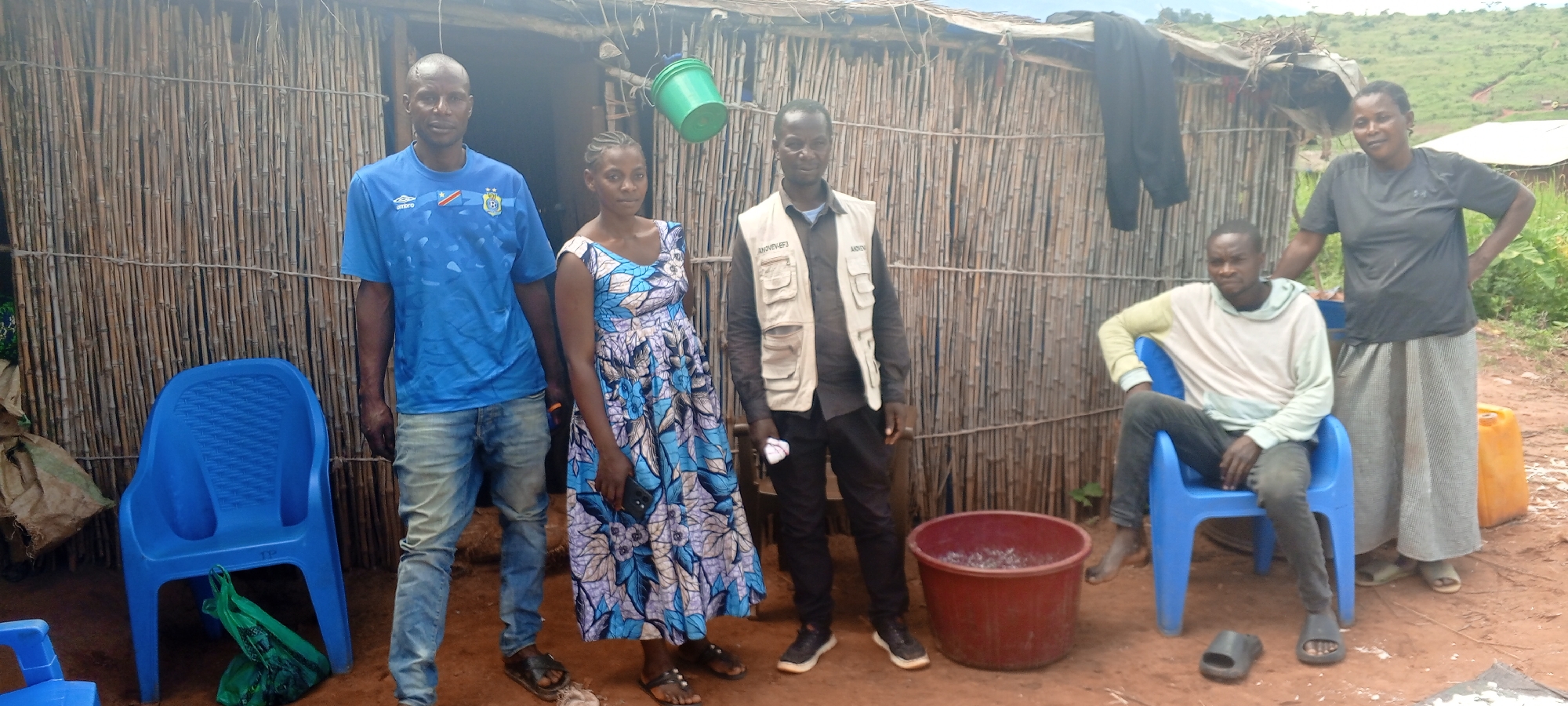 This media file captures five individuals—three men and two women—posing outdoors in Makungu, Democratic Republic of the Congo. They are positioned in front of a rudimentary dwelling constructed with vertical wooden poles or bamboo forming its walls. The ground is reddish-brown earth, and the background features rolling green hills under an overcast sky, suggesting diffused daylight.
From left to right, a man stands wearing a blue t-shirt emblazoned with "UMBRO" and an emblem resembling the Democratic Republic of Congo national football crest. He also wears light-wash jeans and dark sandals. Beside him, a woman in a blue and white floral dress holds a mobile phone and wears sandals. Next to her, another man stands, dressed in a dark long-sleeved shirt covered by a light-colored utility vest, dark pants, and black sneakers. The vest displays text "ANOVEV-EF3" on one pocket and "ANOVEV" on another.
To the right, a man is seated in a blue plastic chair, wearing a light-colored hoodie and dark pants, with black slide sandals placed on the ground in front of him. Behind him, a second woman stands, wearing a dark t-shirt and a light-colored long skirt, also in sandals.
Various objects are visible around the group: an empty blue plastic chair with a green plastic bag underneath it on the left, a red plastic basin filled with small, shiny objects (possibly fish or seeds) on the ground near the center, and a yellow jerrycan on the far right. A green plastic bucket hangs from the dwelling's structure, and dark clothing is draped over its roofline. The individuals generally maintain neutral to slightly serious expressions.