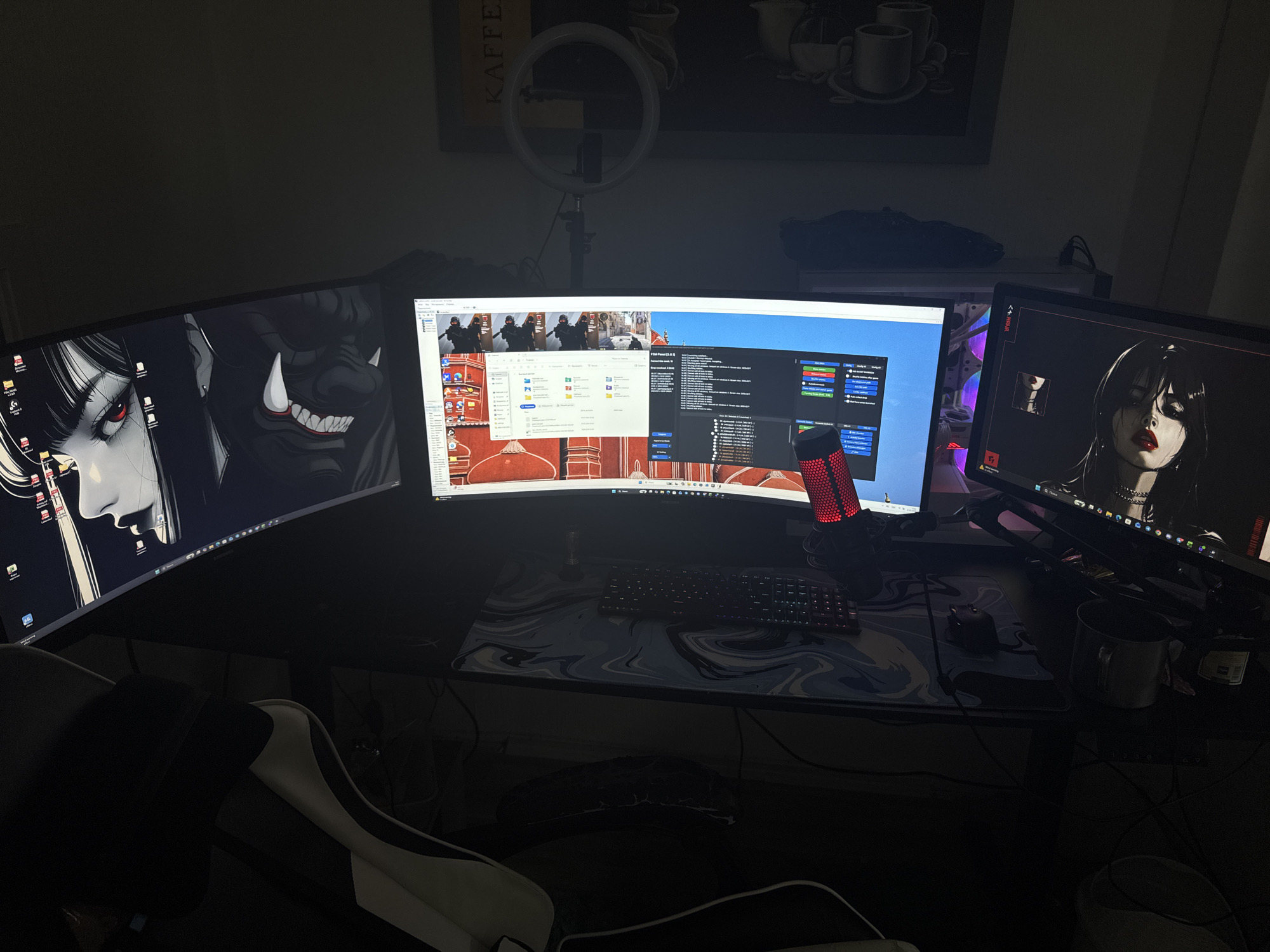 The image depicts a dimly lit room, likely a home office or gaming setup, featuring multiple computer monitors and a desk. The central monitor displays what appears to be a streaming software interface or a video conferencing application, with participants visible in the top portion and various windows and controls below. To the left and right are two other monitors, each displaying stylized anime-style artwork. The desk itself is covered by a large mousepad with a marbled design, a keyboard, a mouse, and a red microphone. A gaming chair is partially visible in the foreground. In the background, a ring light on a stand is mounted above the central monitor, suggesting the space is used for content creation. A framed picture with text like "KAFFEE" is mounted on the wall, along with some potted plants. The scene is dark, with artificial light sources primarily illuminating the monitors and the immediate desk area. The city of Neubrandenburg, Germany is not mentioned or indicated in the image.