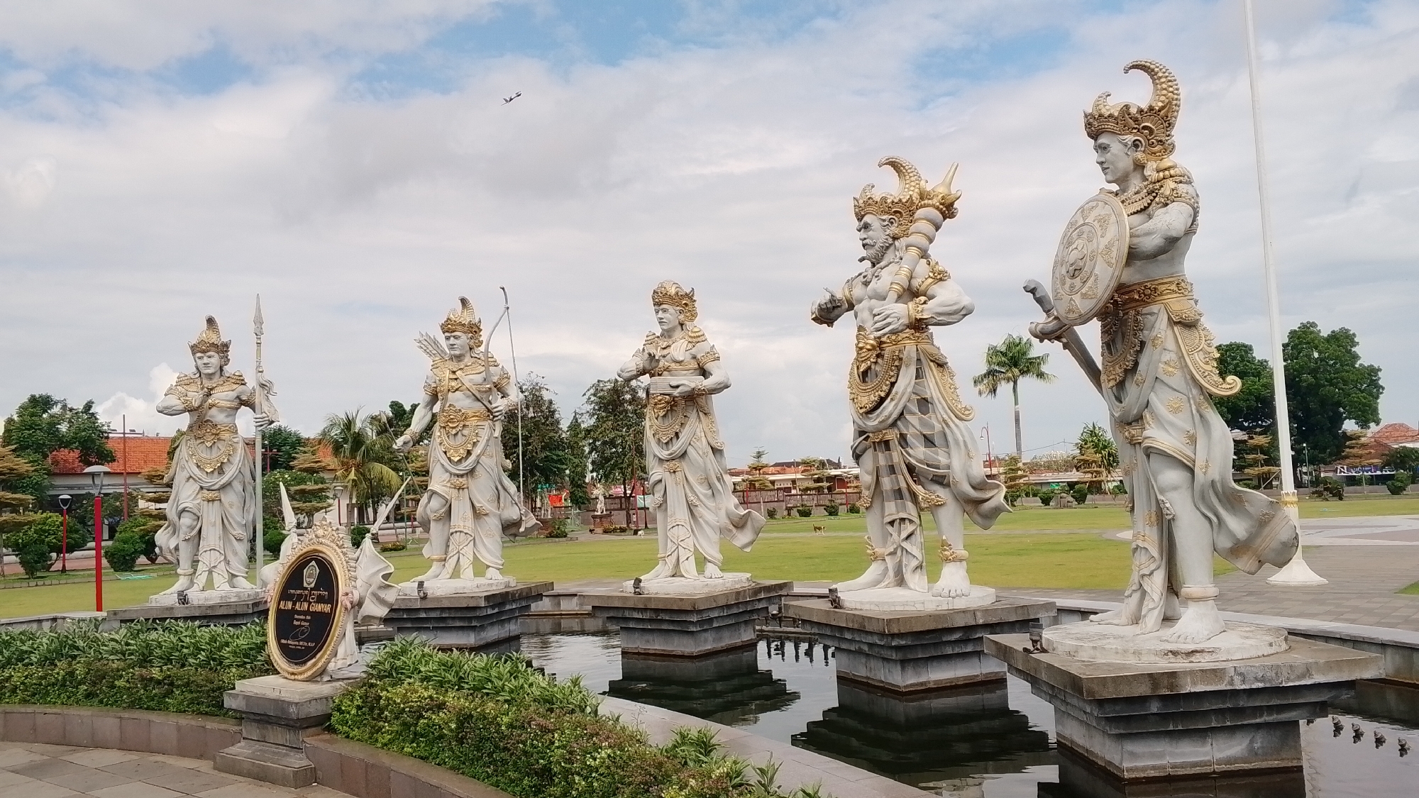 A wide shot captures a row of five large, white statues with gold accents, each adorned with intricate headdresses and traditional Balinese clothing. The statues are positioned in a shallow pool of water, their bases appearing to be made of stone. From left to right, the first statue holds a spear, the second has a bow and arrow, the third holds a bowl or offering, the fourth clutches a mace or staff, and the fifth is depicted with a sword and shield. Behind the statues, a green grassy field stretches out, dotted with trees and some buildings. In the distance, a palm tree is visible, and the sky is partly cloudy with a small airplane in the upper left quadrant. A sign with gold lettering and an emblem is placed in front of the first two statues. The overall scene appears to be an outdoor monument or plaza.