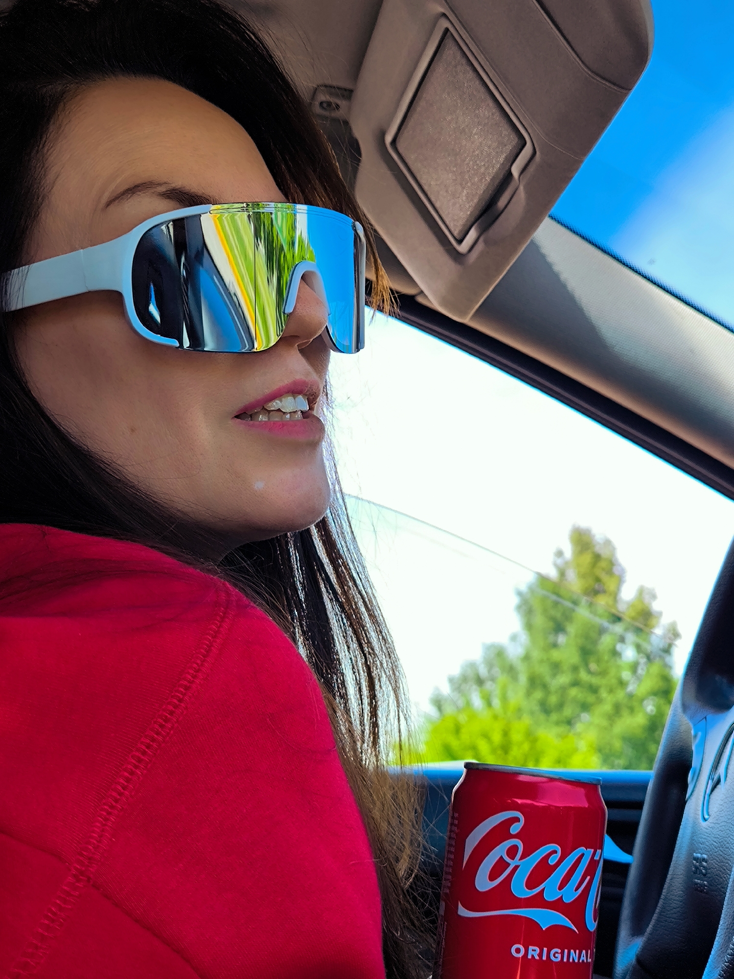 The image captures a woman in the driver's seat of a car. She is wearing reflective sunglasses with a light-colored frame and a red hooded sweatshirt. She has dark hair and pink lipstick. A can of Coca-Cola Original is visible near the steering wheel. The scene is presumably inside the car, with a sun visor and the car's interior visible. The background outside the car features green trees and a bright blue sky, suggesting an outdoor setting. The location is not identifiable as Riga, Latvia based on the information provided.