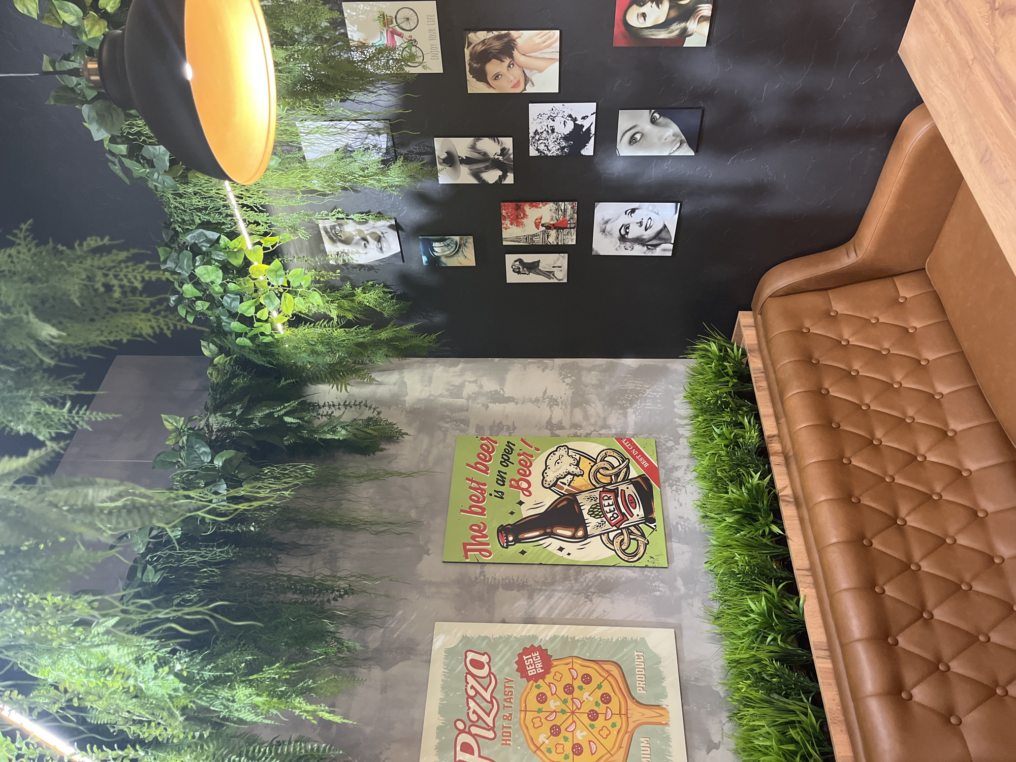 This image shows a scene inside what appears to be a restaurant or cafe. The walls are decorated with a combination of framed pictures, posters, and greenery.
On the top wall, which is a dark gray color, there are several framed pictures. Most of these appear to be black and white portraits or scenes, while one has a bicycle, and another has the Eiffel Tower.
The lower wall is light gray. Two posters are visible on this wall. One reads "The best beer is an open Beer!" with an illustration of a beer bottle and pretzels. The other poster features a pizza and the words "Pizza Hot & Tasty Best Price".
To the left of the wall is an abundance of artificial greenery, creating a garden-like effect. A black and gold pendant light hangs from the ceiling.
To the right of the wall is a brown, tufted sofa, with more artificial greenery positioned along the edge of the sofa.
The location is not identifiable from the provided image.