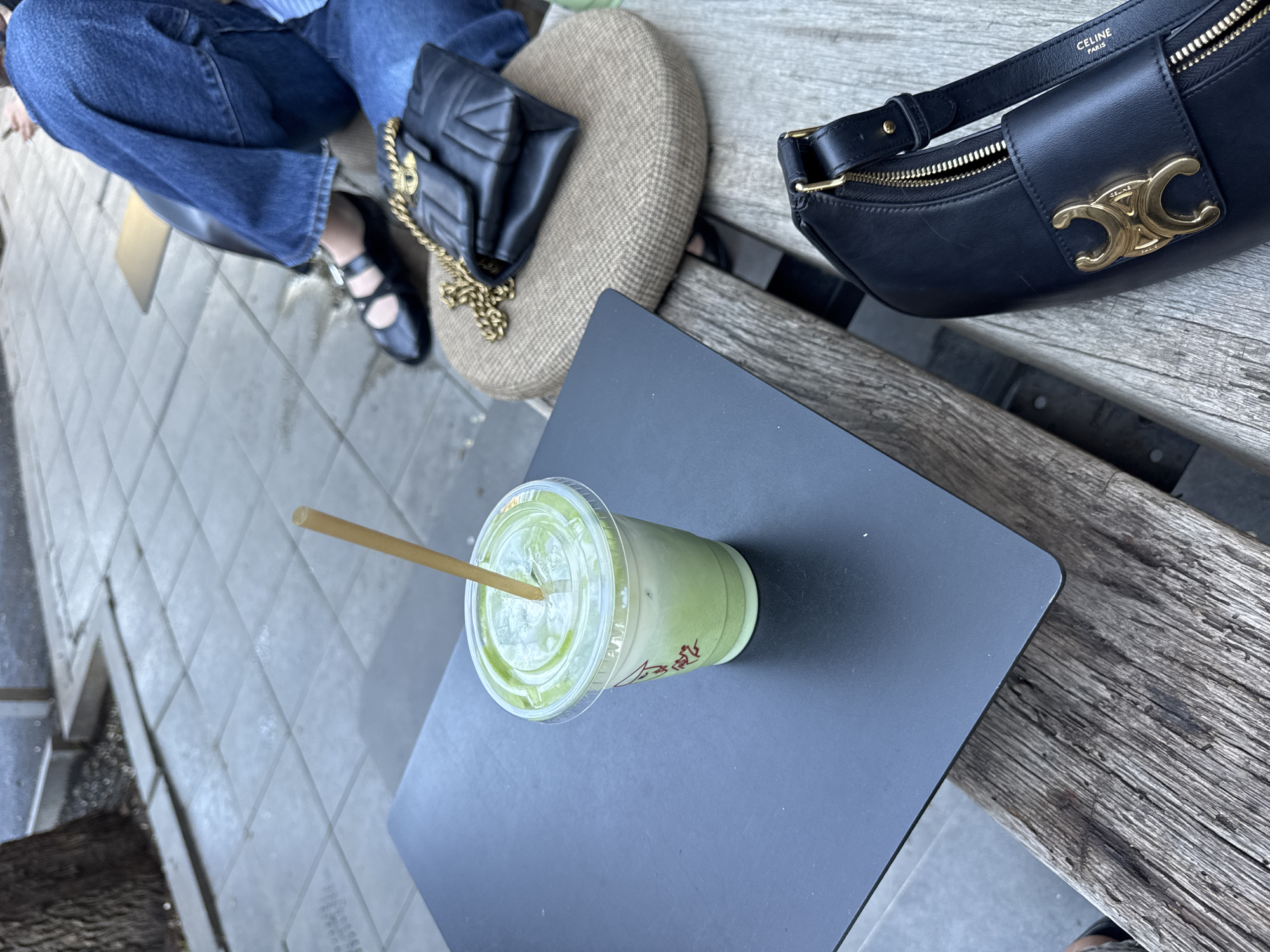 A person wearing blue jeans and black shoes sits on a bench beside a dark grey metal table. On the table is a clear plastic cup filled with a green liquid, presumably a matcha latte, and a brown paper straw. A black Celine bag is placed on the bench, near the table. Next to the person is a small, round cushion with a black quilted purse placed on it. The scene takes place outdoors, likely in an urban area. The location is the city of Yerevan, Armenia.