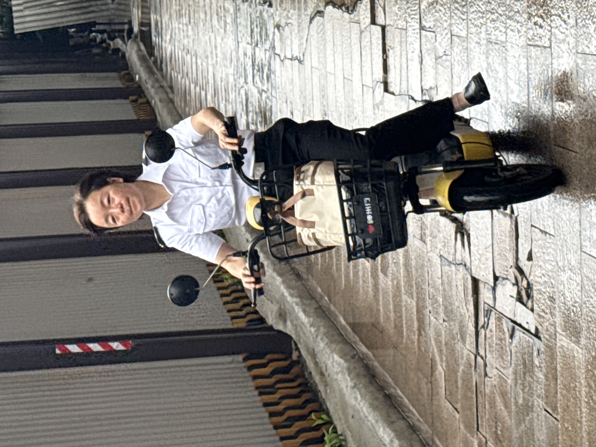 A woman is riding an electric scooter on a wet, paved surface in Nan'an District, China. The woman is wearing a white, button-down shirt and black pants, and black flat shoes. The scooter is yellow and black, with a black basket on the front containing a tan bag. The scooter has black rearview mirrors. The surface she is riding on is wet and reflects light. The background includes a wall with a red and white striped marker and a textured barrier. The surface appears to be made of rectangular gray paving stones.