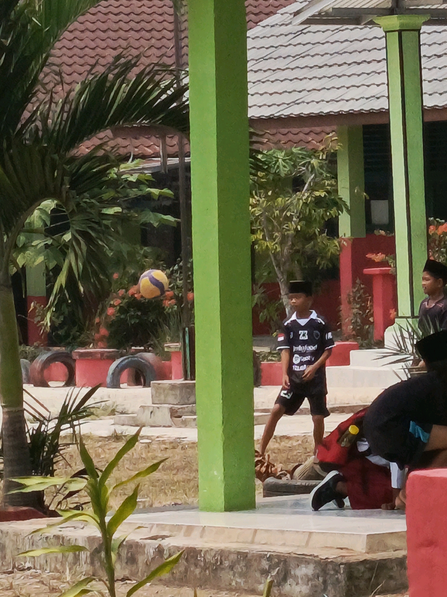 The image depicts an outdoor scene, likely in a schoolyard or park, with a group of children present. In the foreground, a bright green pillar stands prominently. To the left, a palm tree with large fronds is visible, partially obscuring a building with a tiled roof. A yellow and blue volleyball is suspended in the air mid-game.
In the mid-ground, several children are engaged in an activity. One boy, wearing a black and white jersey with the number 23 and a black peci (a cap common in Indonesia), stands looking to his left. Another boy, also wearing a peci, is partially visible to the right. In the foreground, another child is hunched over or lying down, wearing a black shirt and white shoes.
The environment includes red planters, some with tires repurposed as decorative elements. The ground is dry and dusty. The background reveals more of the building's structure and greenery. The scene appears to be illuminated by bright daylight, suggesting it is daytime. The overall atmosphere suggests a casual, playful moment among children.