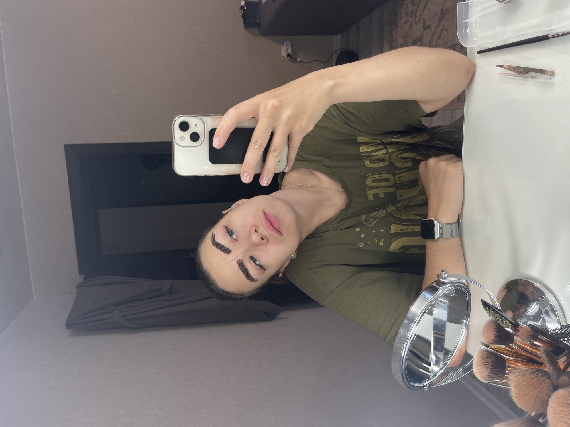 A young woman with dark brown, freshly dyed eyebrows and dark eyeliner is taking a selfie in a mirror. She is wearing a dark green t-shirt and a silver smartwatch on her left wrist. She is sitting at a white table with a silver makeup mirror and a collection of makeup brushes in front of her. Tweezers and a small brush are also visible on the table. The background shows a wall with a gray curtain and a doorframe. The scene appears to be in a residential setting, likely a bathroom or dressing area. The image does not provide any information about the location in Chaikovsky, Russia.