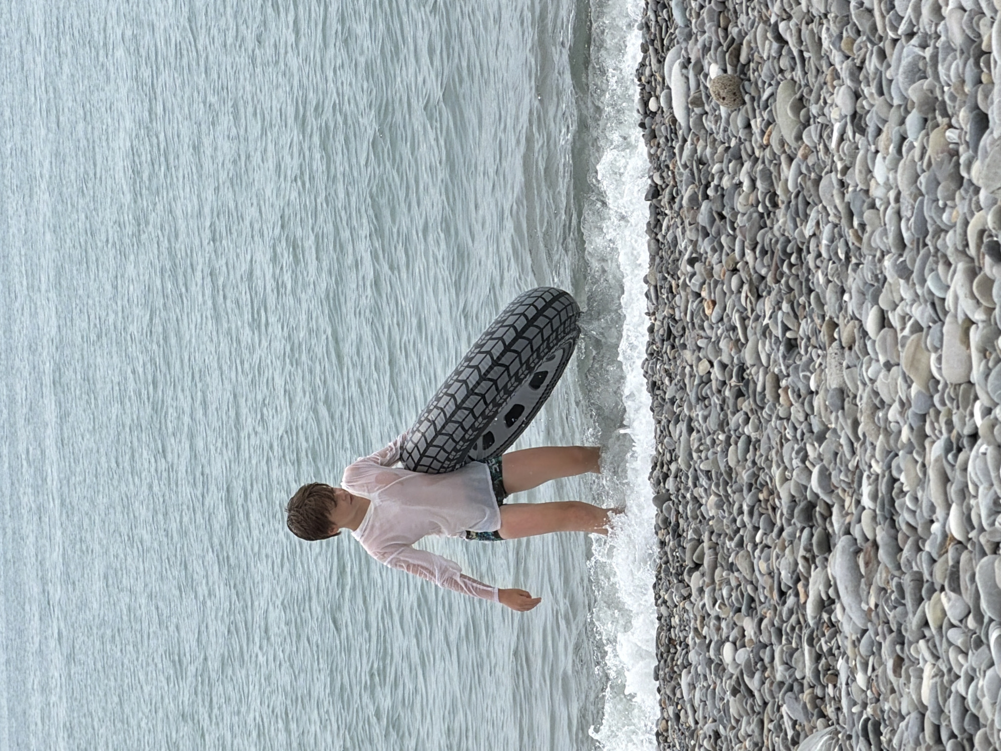 The image depicts a young individual, appearing to be a boy, standing in shallow water at the edge of a pebble beach. He is wearing a translucent white long-sleeved shirt and patterned swim shorts. He is holding a large, black inflatable tire with a tire tread pattern. The water is calm with small waves breaking on the shore. The beach is covered in smooth, grey pebbles. The background consists of a body of water, likely the Black Sea, under an overcast sky. The scene is consistent with a casual day at the beach in Sochi, Russia.
