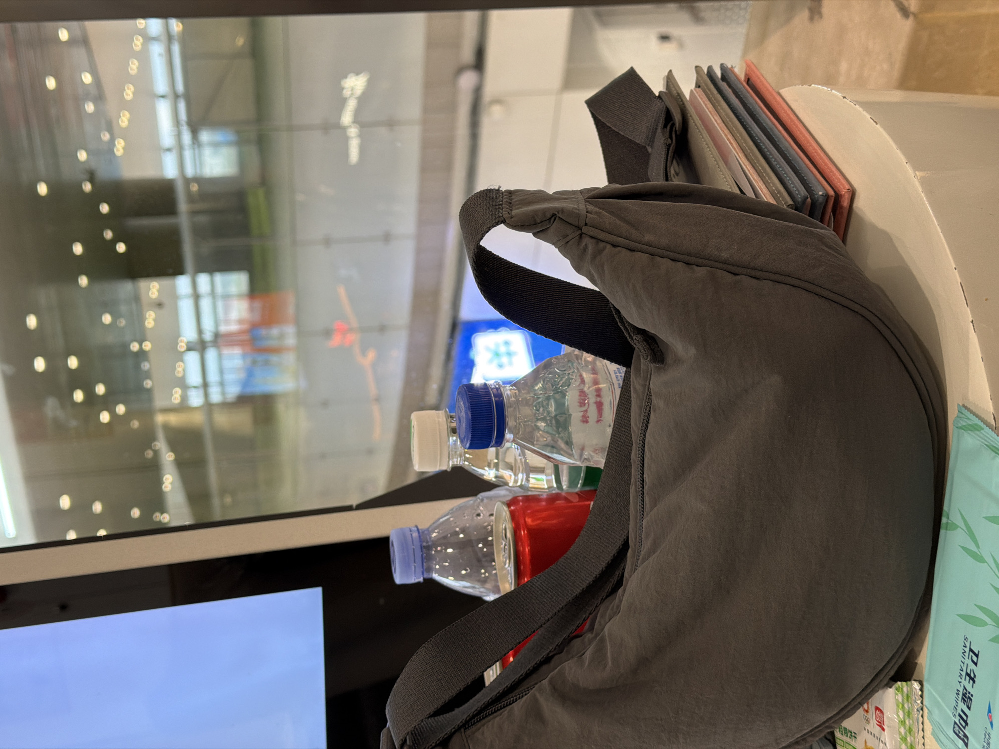 A dark gray crossbody bag is positioned on a cream-colored cylindrical surface. To the left of the bag, there are two bottled beverages and a red can. Behind the bag, a large window reflects lights and an indoor environment with a railing. To the right of the bag, a stack of colorful booklets or folders is visible. In the foreground, partially visible, are two packets of wet wipes. The overall setting appears to be an indoor public space, possibly a waiting area or a shop, in Guangzhou City, China, suggested by the packaging of the wet wipes.