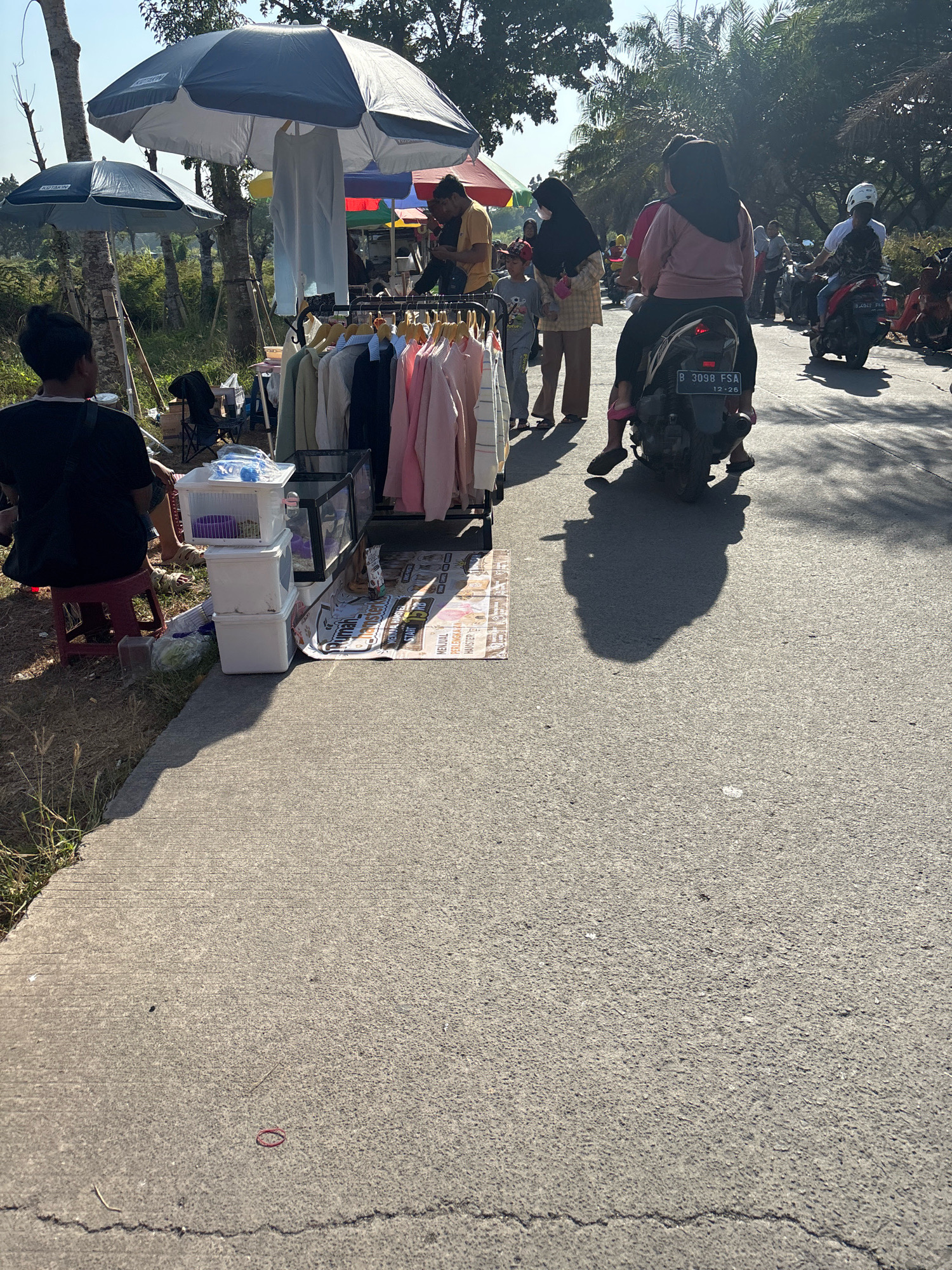 In Telagamurni, Indonesia, a street-side market is captured on a sunny day. A person sits on a red stool to the left, facing a display of clothing on a rack. The clothing consists of various tops, including t-shirts and blouses, in pastel colors and neutral tones. To the right of the clothing rack are stacked white storage containers. Behind the market stall, several people are visible, some standing and others on motorcycles. One motorcycle with two occupants, a driver and a passenger wearing a black headscarf and pink top, is in motion on the paved road. Another motorcycle with a driver wearing a white helmet is also visible further down the road. Trees and foliage line the background, suggesting a semi-rural or suburban setting. The ground is a mix of concrete walkway and asphalt road. A red rubber band lies on the concrete walkway in the foreground.